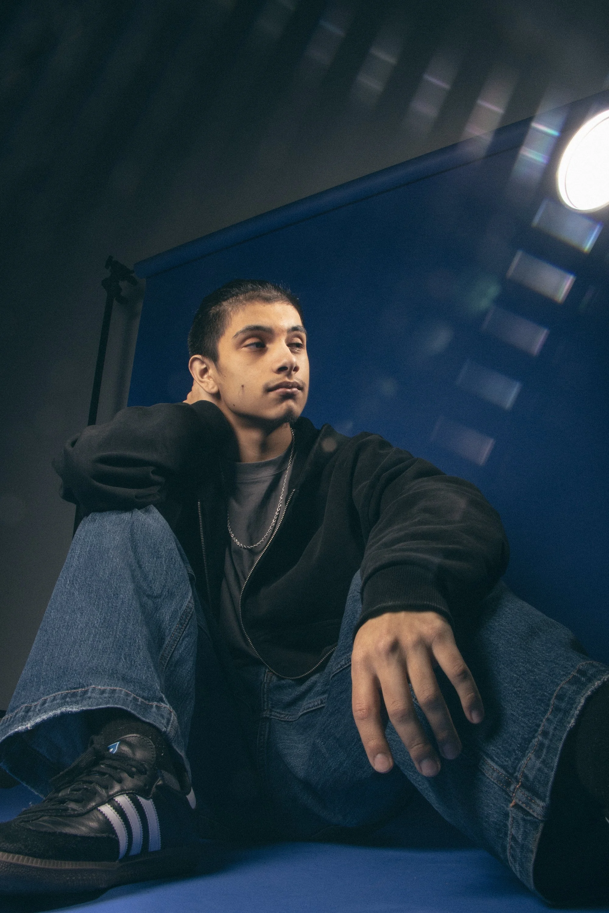 A young man sitting on the floor in a photography studio with a blue backdrop and professional lighting equipment overhead. He is wearing a black jacket, gray shirt, jeans, and sneakers, and is resting one arm on his knee with his hand relaxed.