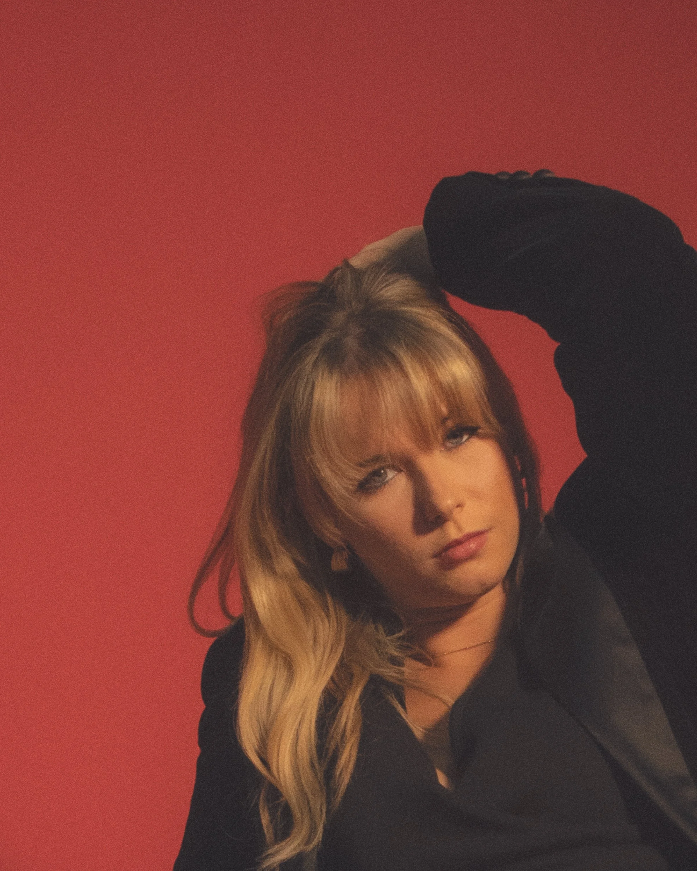 A woman with long, blonde hair, wearing a black top, rests her arm on her head. She is standing in front of a solid red background.