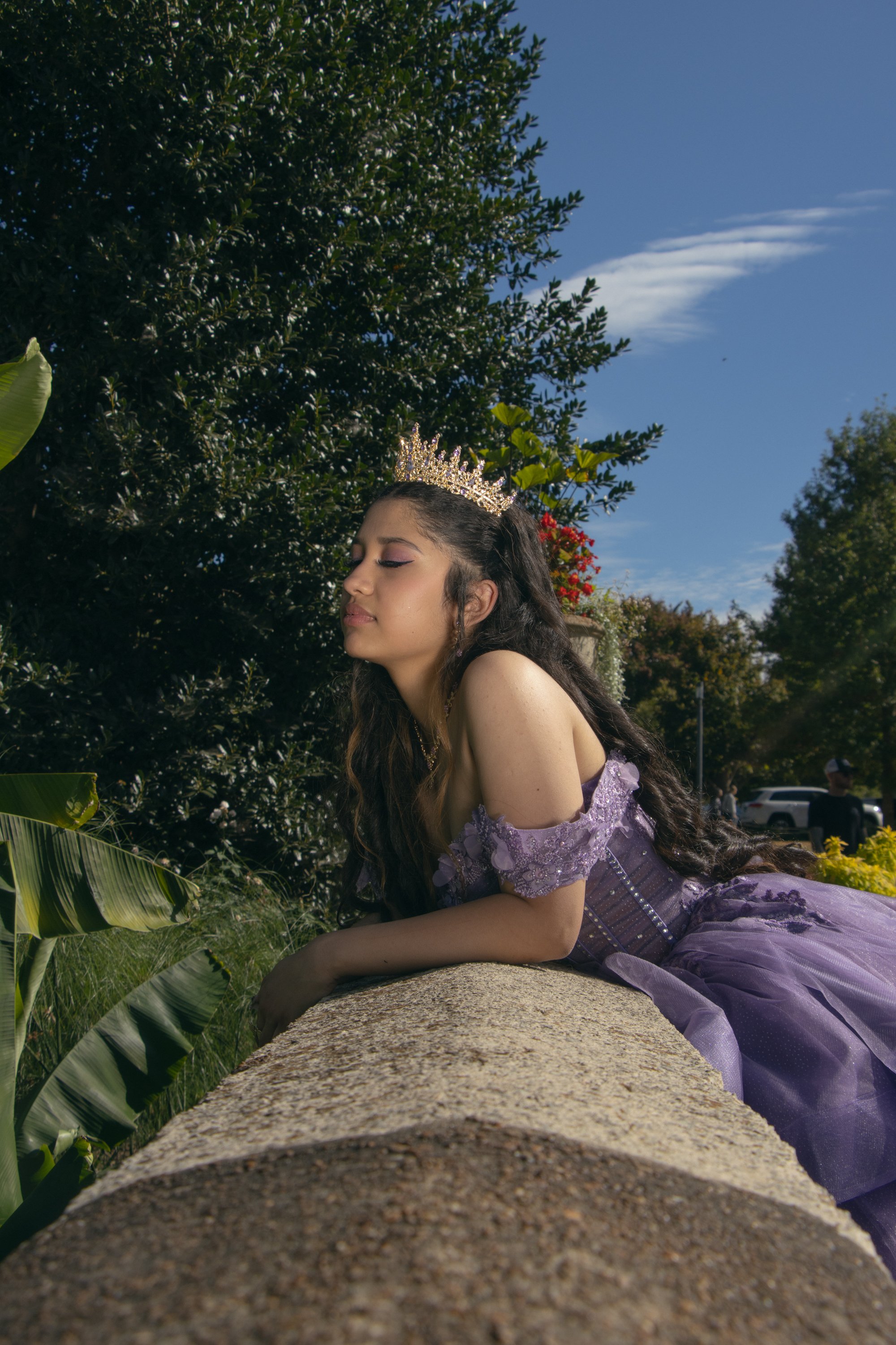 Young girl wearing a lavender dress and a tiara, lying down on a stone ledge outdoors with lush green trees and a partly cloudy blue sky in the background.