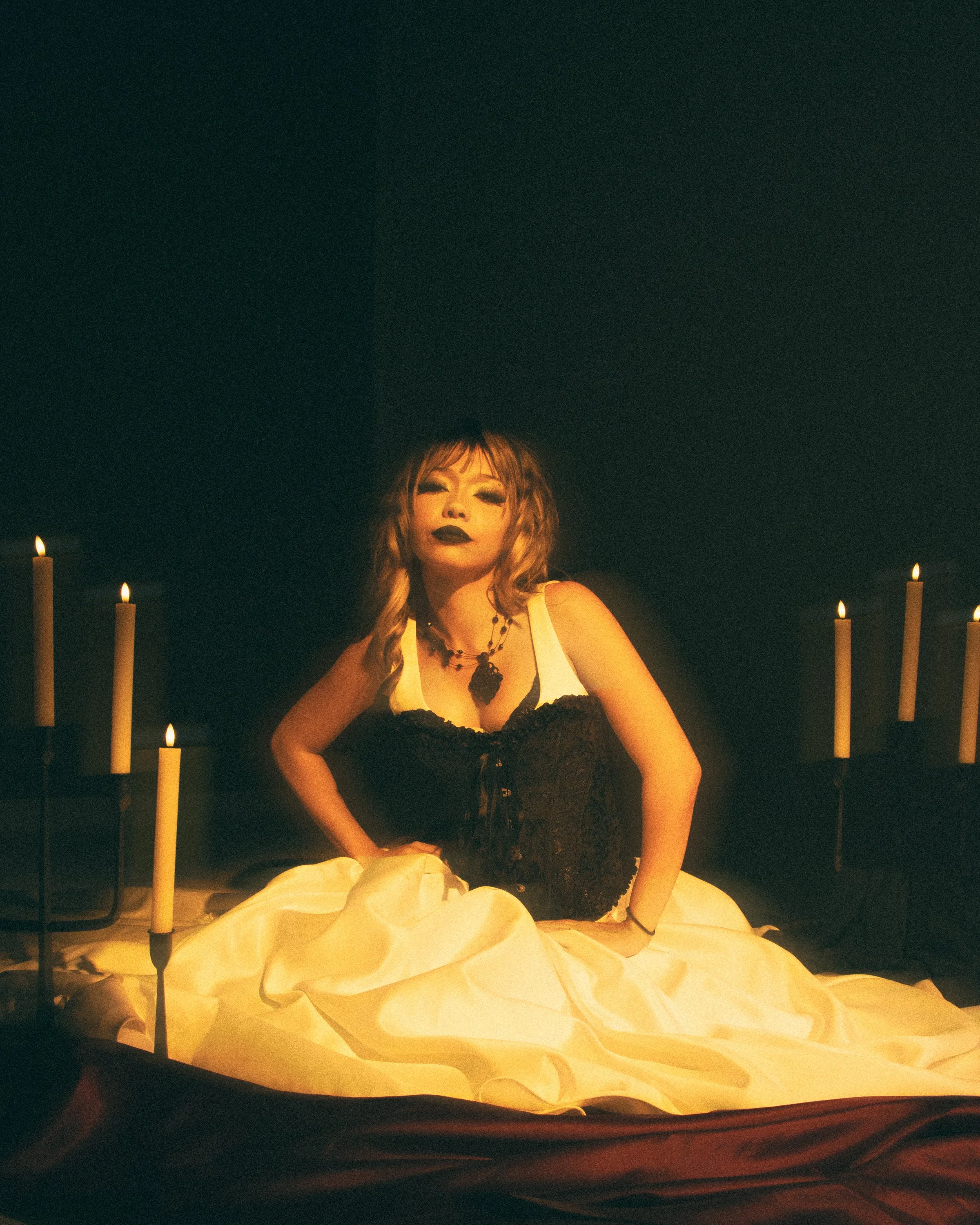 A woman with long wavy hair, dark makeup, and gothic style clothing sitting among candles in a dimly lit room.