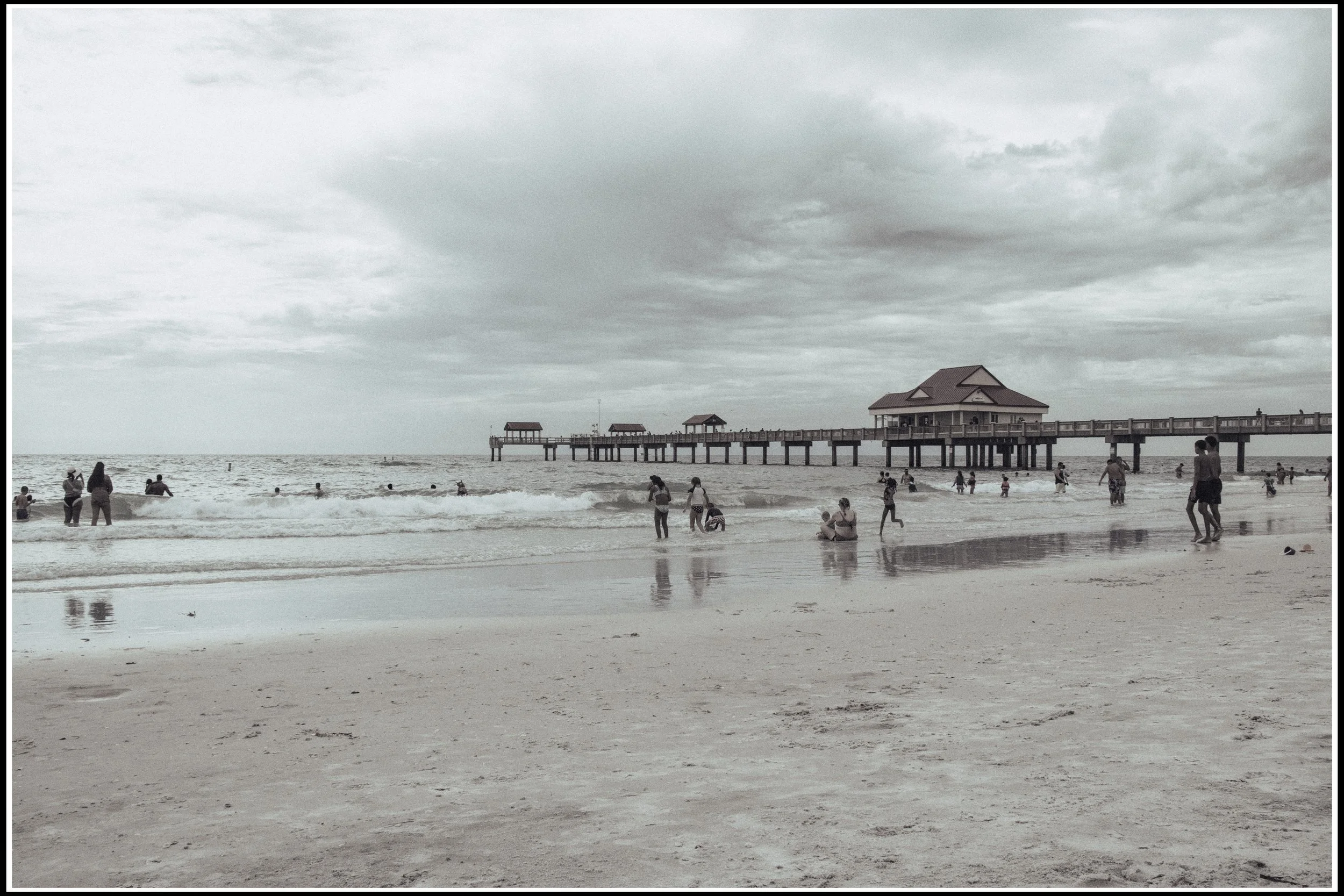 People enjoying a cloudy day at the beach; some are walking, swimming, and sitting on the sand near a pier with small pavilions and a large house at the end.