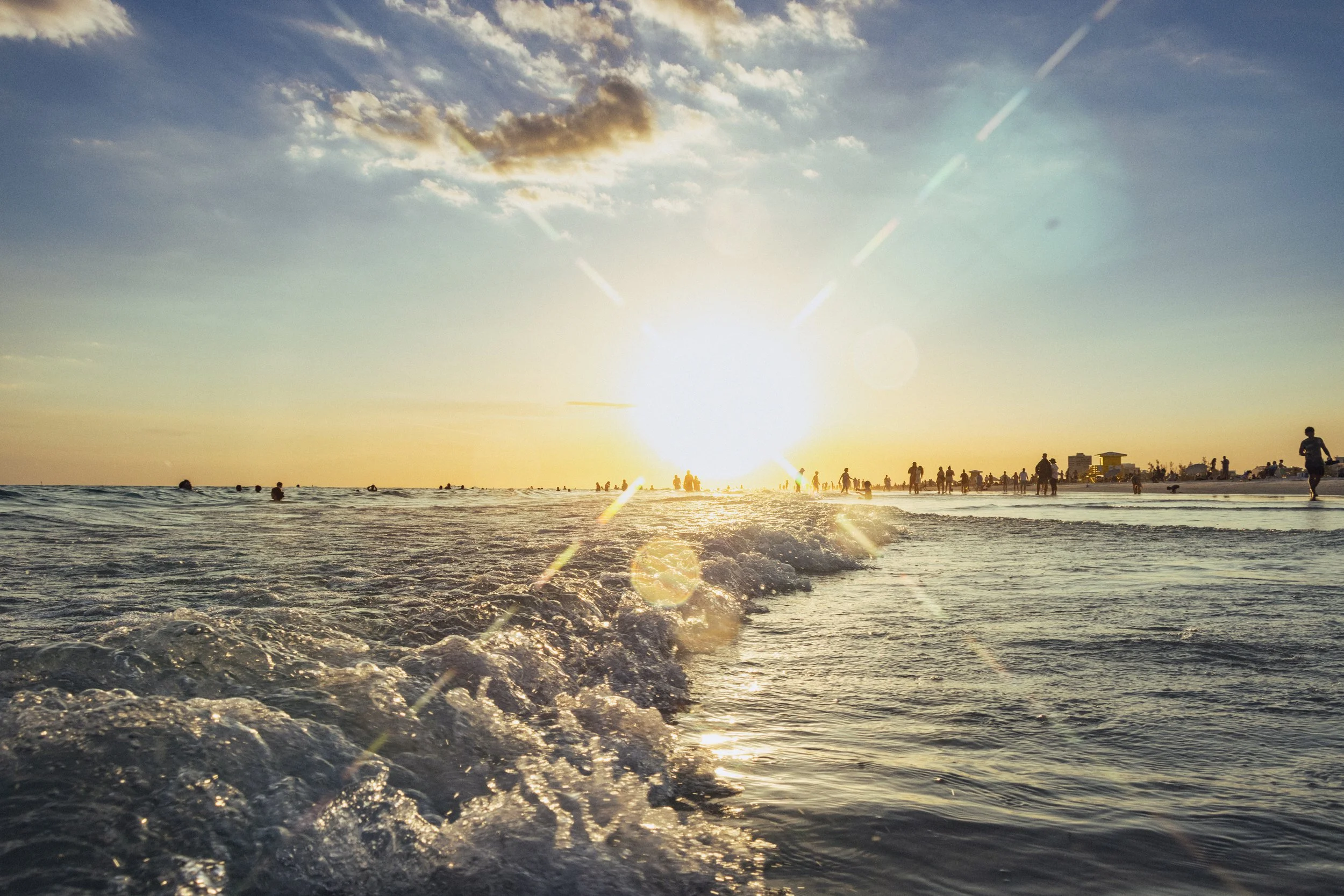 Beach scene at sunset with people swimming in the water and walking along the shore, city buildings in the distance, and the sun low in the sky with lens flare.
