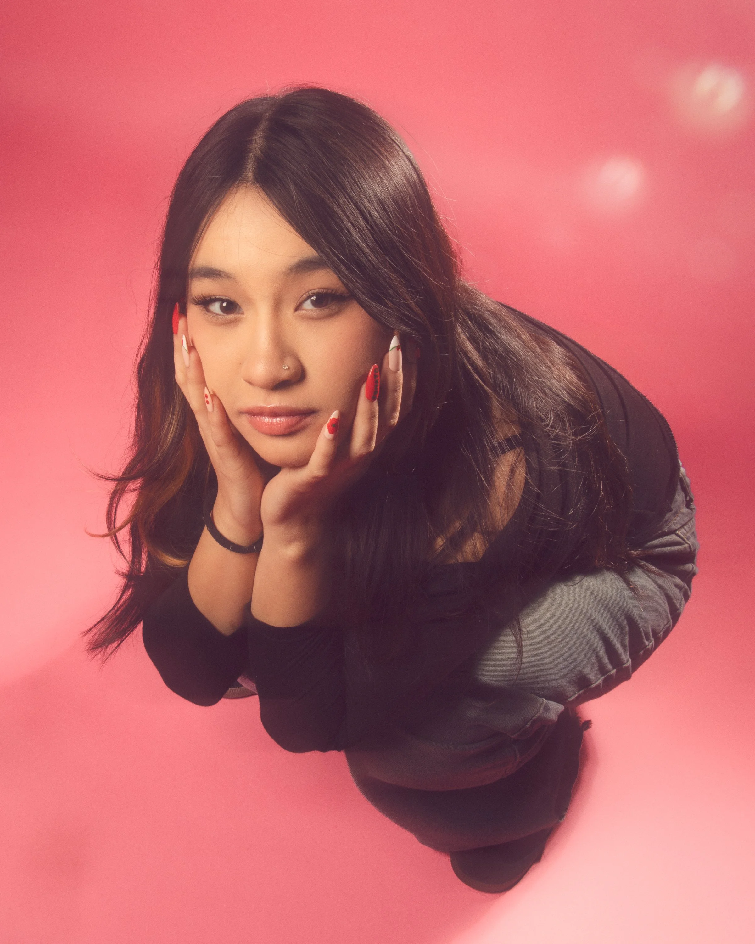 A young woman with long dark hair, wearing makeup, holding her face with both hands, looking directly at the camera, against a pink background.