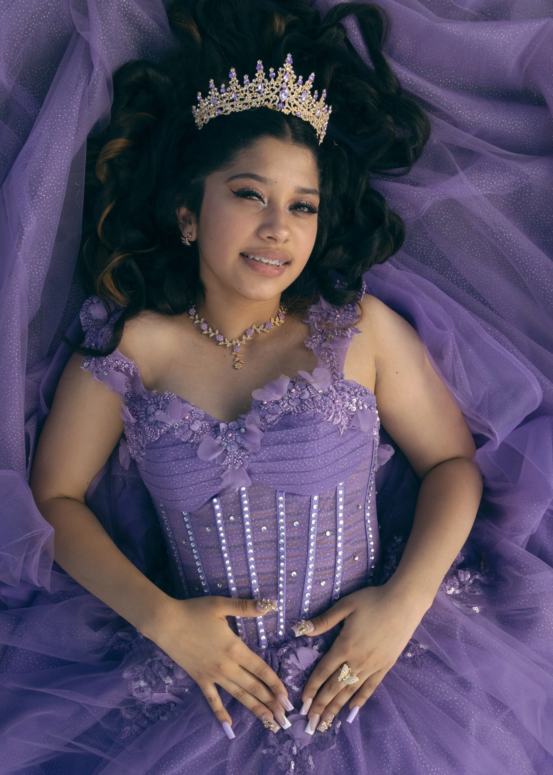 A woman dressed in a purple gown with lace and bead embellishments, wearing a silver tiara with purple gemstones, and jewelry including earrings, a necklace, and rings. She has long dark wavy hair, makeup with winged eyeliner, and leopard-print nails