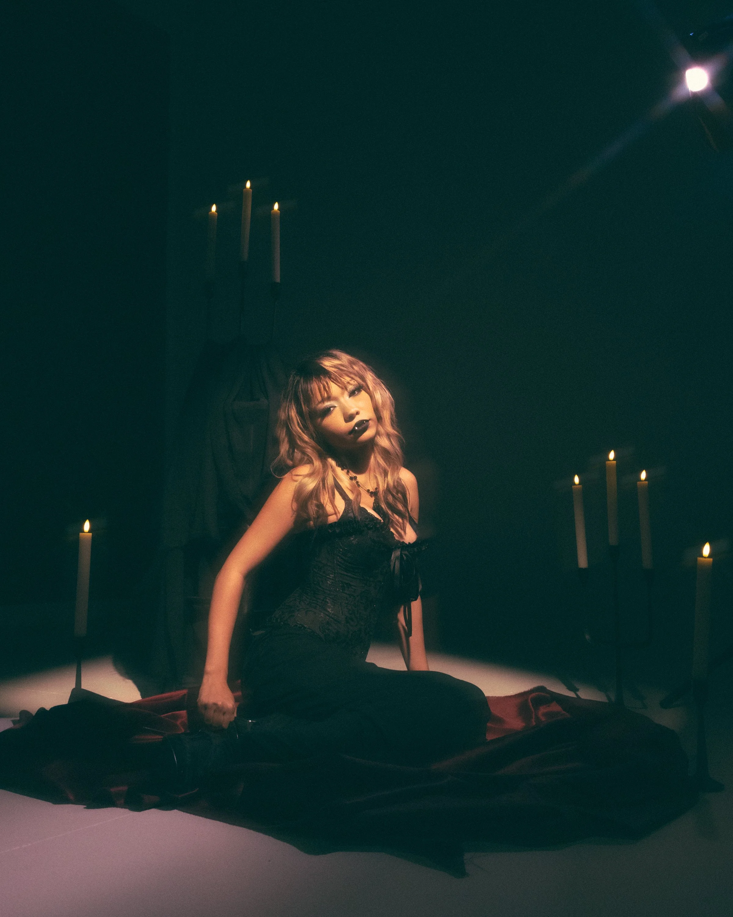 Dark room with candles and a woman with pink hair in gothic attire sitting on a platform, illuminated softly.