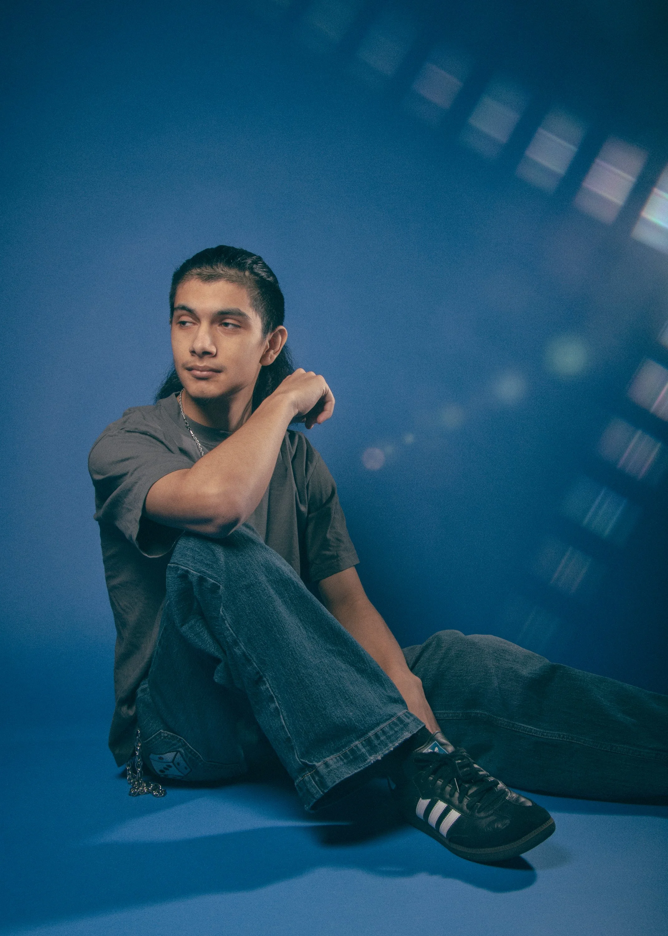 A young man with long black hair seated on the floor against a blue background, wearing a gray t-shirt, black Adidas sneakers, and blue jeans. He has a chain hanging from his pocket and is looking to his right with a slight smile.