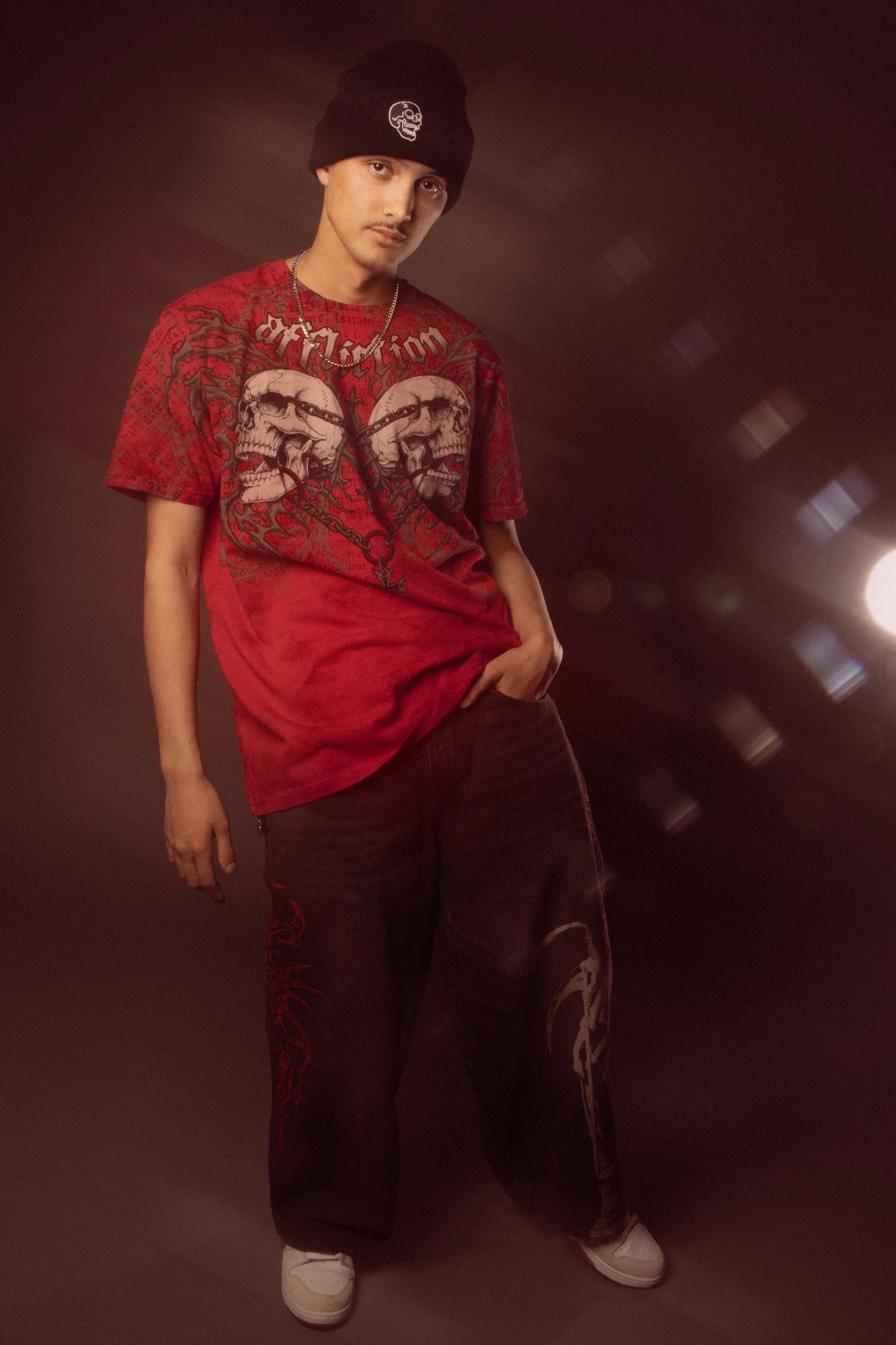 Young man wearing a black beanie with a skull logo, red graphic t-shirt with skull and chain design, black cargo pants with red embroidery, and white sneakers, standing in a dimly lit room.