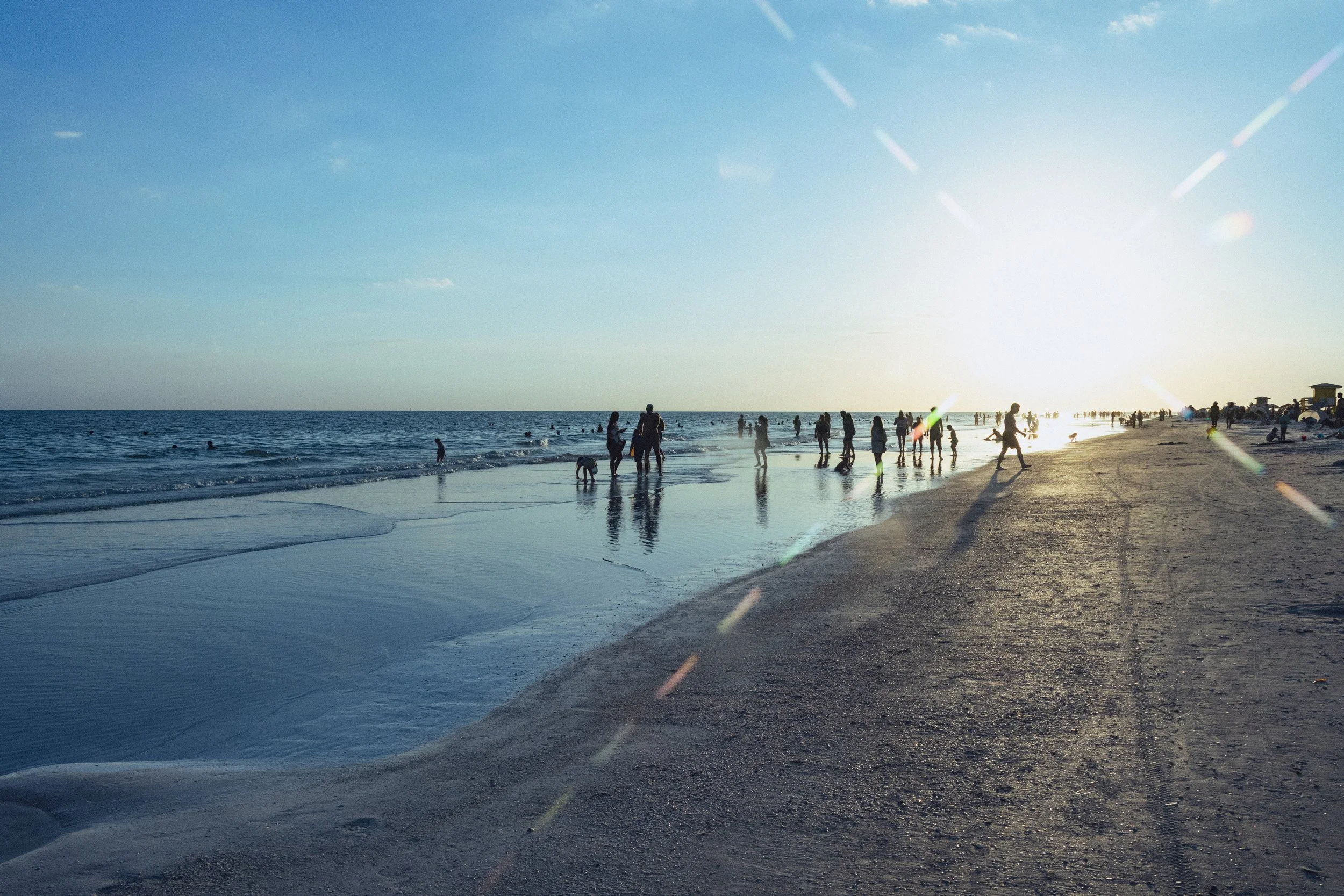 People enjoying a beach scene during sunset or late afternoon with the sun low in the sky, reflecting on the water, and lifeguard stands on the shoreline.