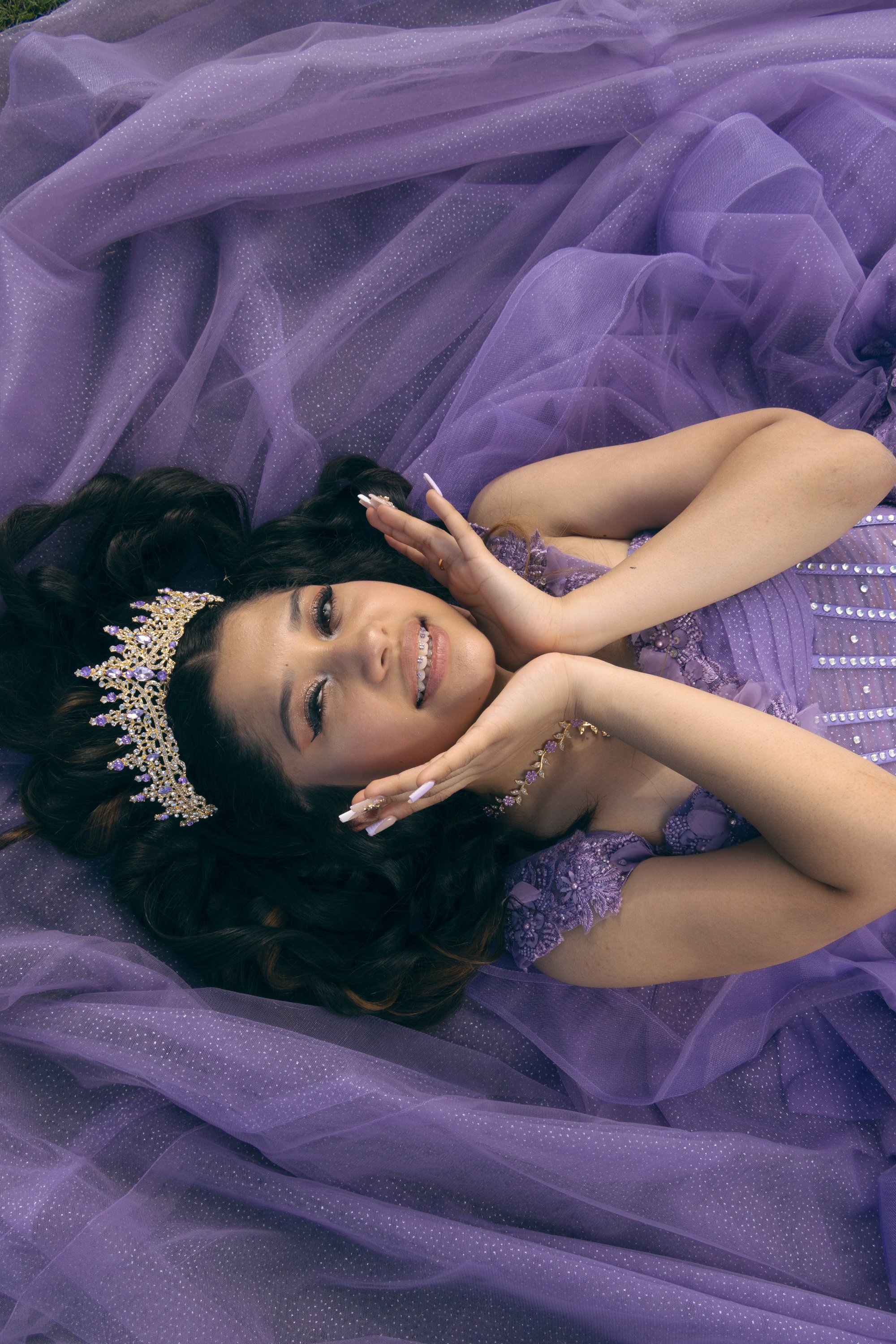 Young woman in a purple princess dress lying down on purple tulle fabric, wearing a golden tiara with purple and clear gemstones, smiling with her hands near her face.