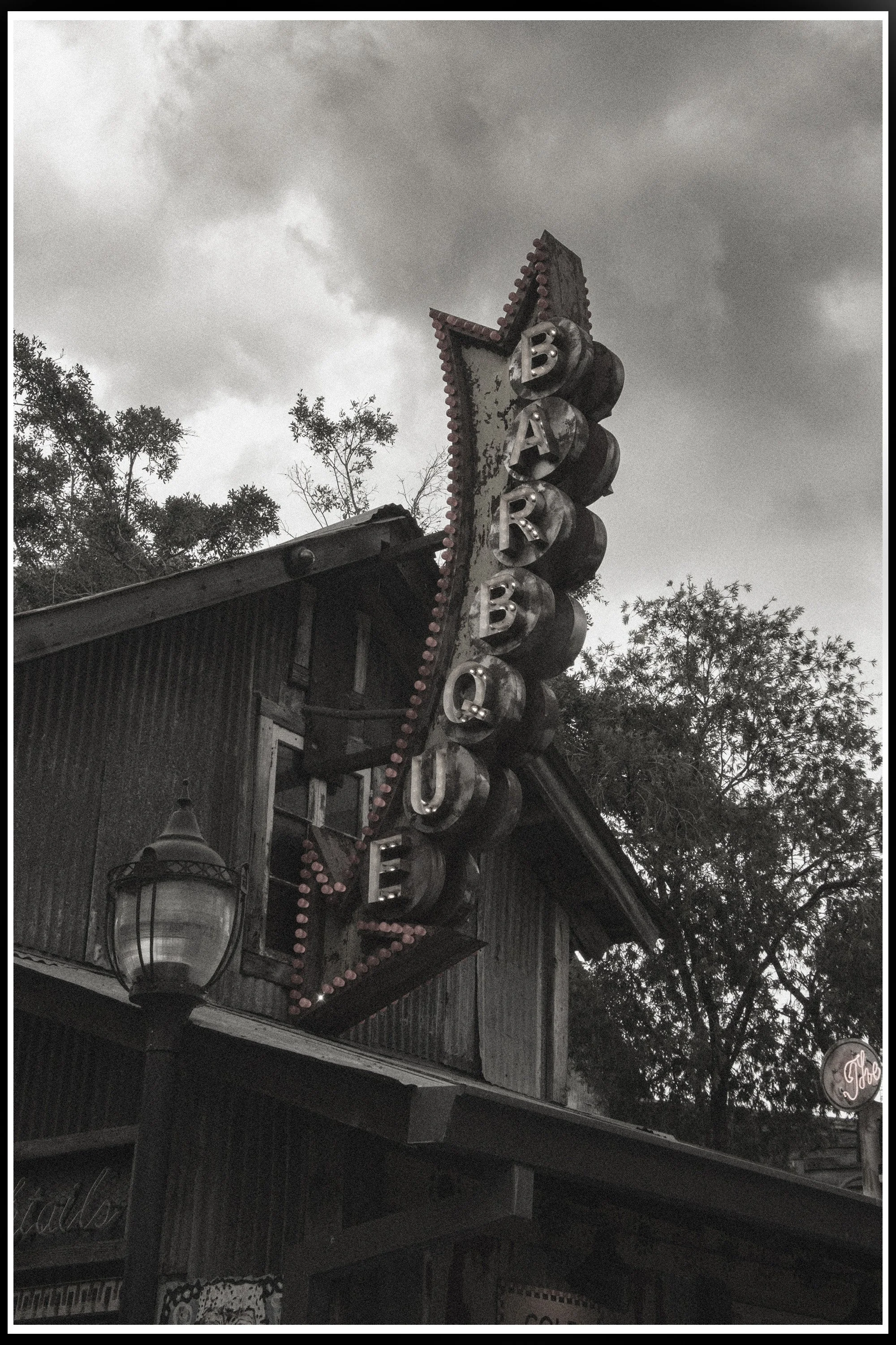 Old vintage wooden sign with parts falling off, displaying the word 'Bar' vertically with large lightbulb bulbs around it, attached to a rustic building exterior with a vintage lantern and trees in the background, under cloudy sky.