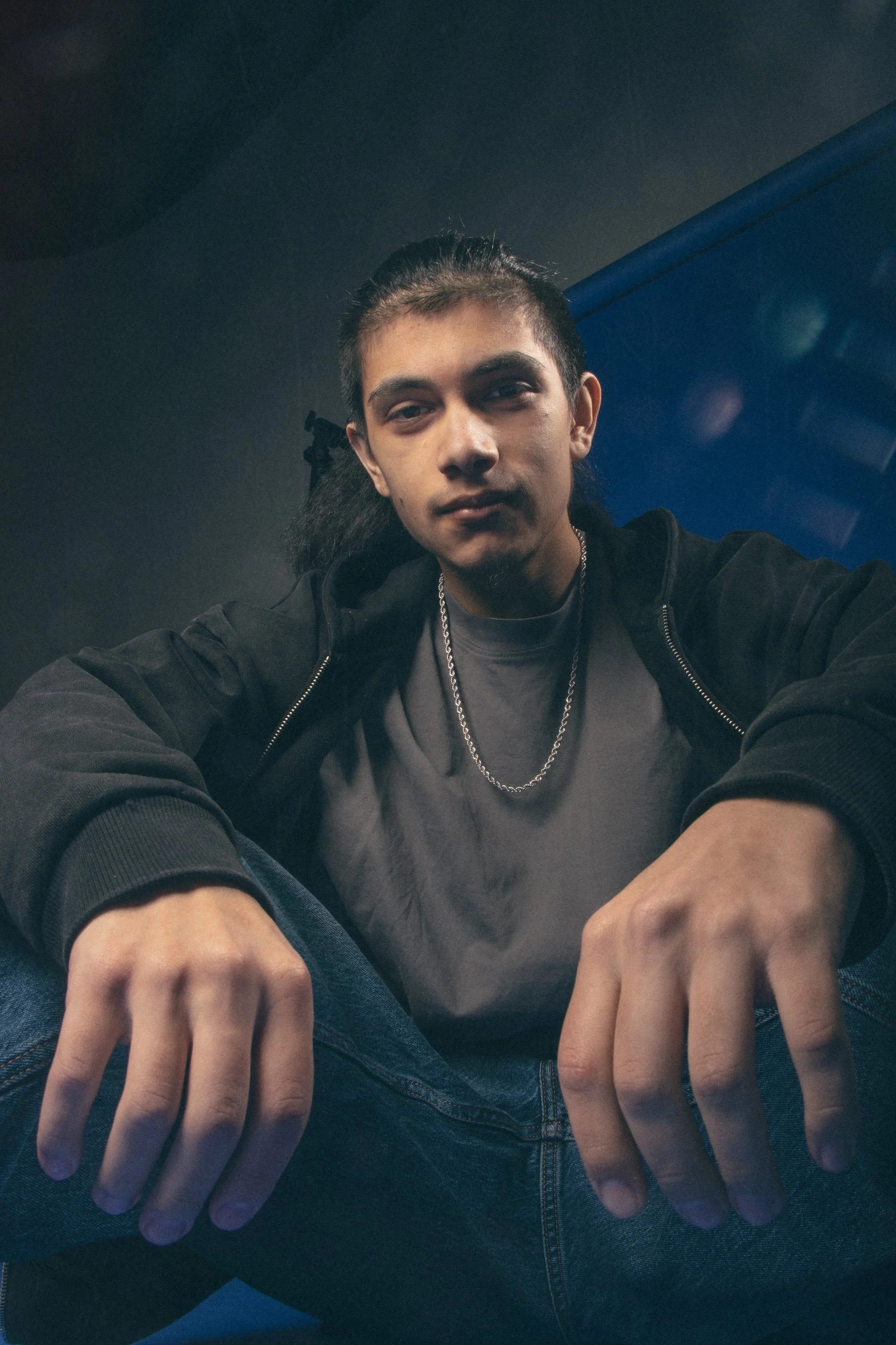 Close-up of a young man with long dark hair, wearing a black jacket, gray T-shirt, and silver chain, sitting with hands resting on knees, in front of a dark background with blue elements.