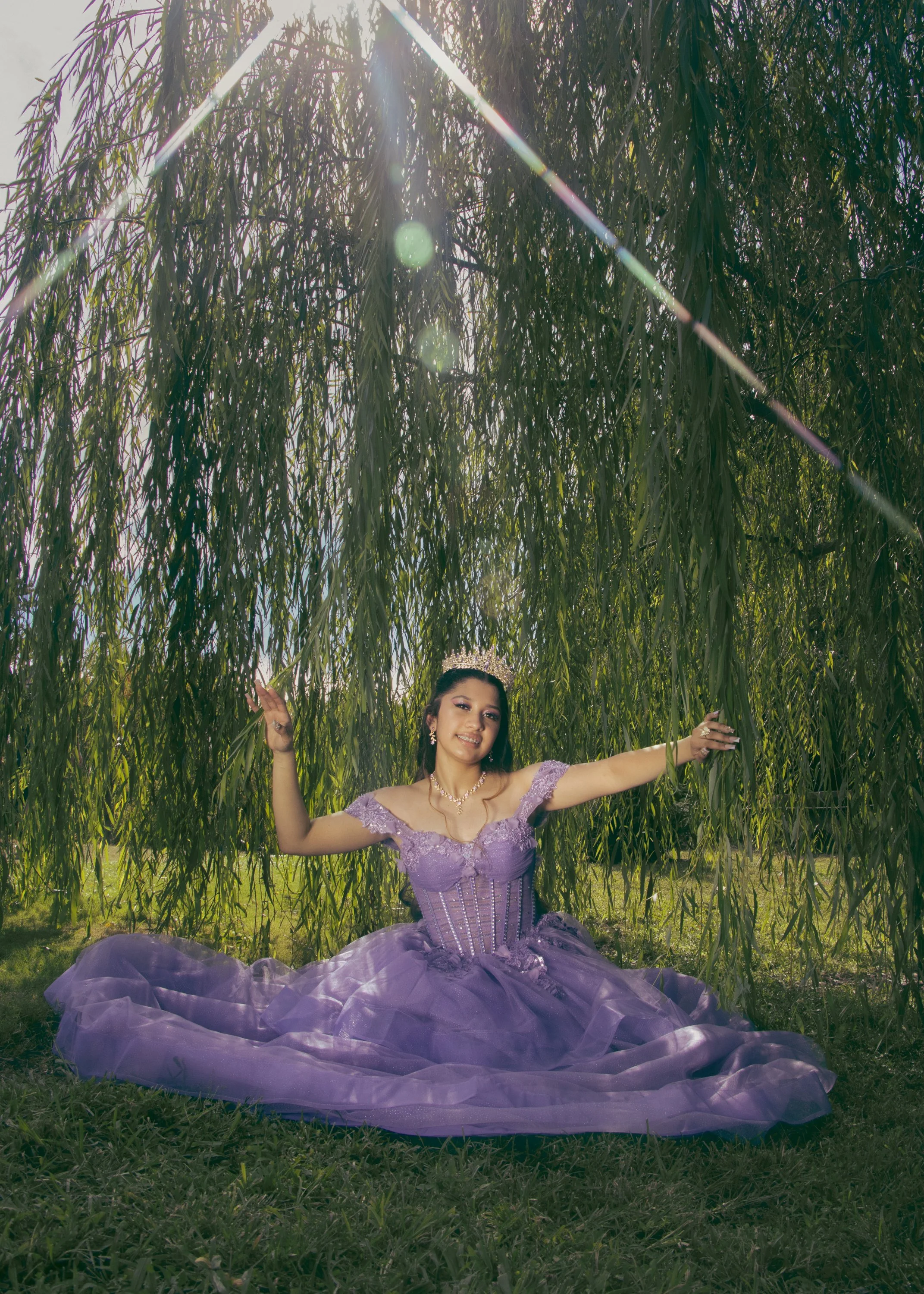 A woman dressed in a purple gown sitting on grass under a tree with hanging branches, smiling, with sunlight coming through the leaves.