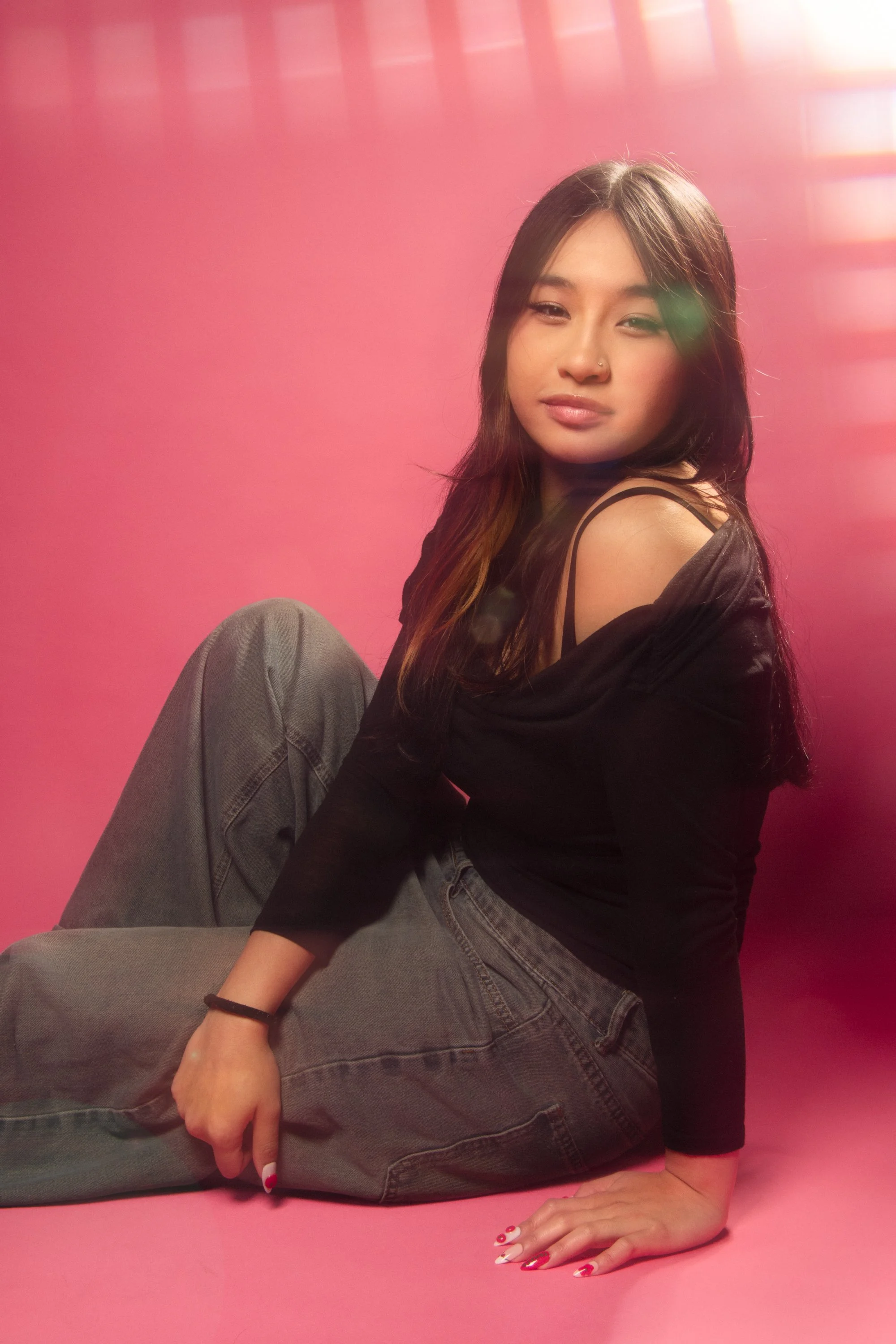 Young woman with long dark hair and a nose piercing sitting on a pink surface in front of a pink background, wearing a black top with shoulder straps, gray jeans, with her right hand on the ground and her left arm resting on her knee.