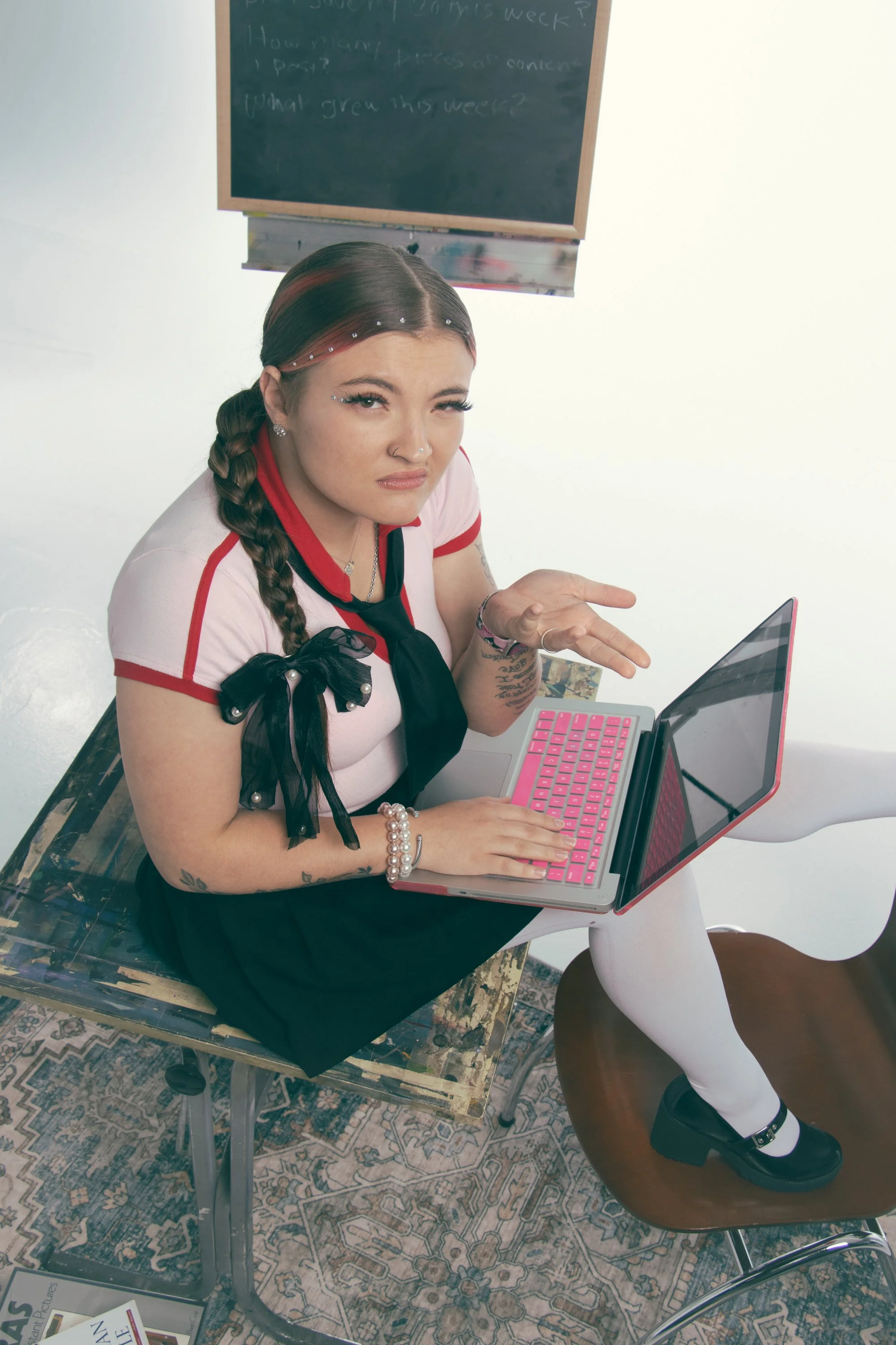 A woman sitting on a desk with a laptop on her lap, making a confused or skeptical expression. She has multiple piercings and tattoos, and is dressed in a pink and red shirt with a black bow. Behind her is a chalkboard with writing, and the scene inc