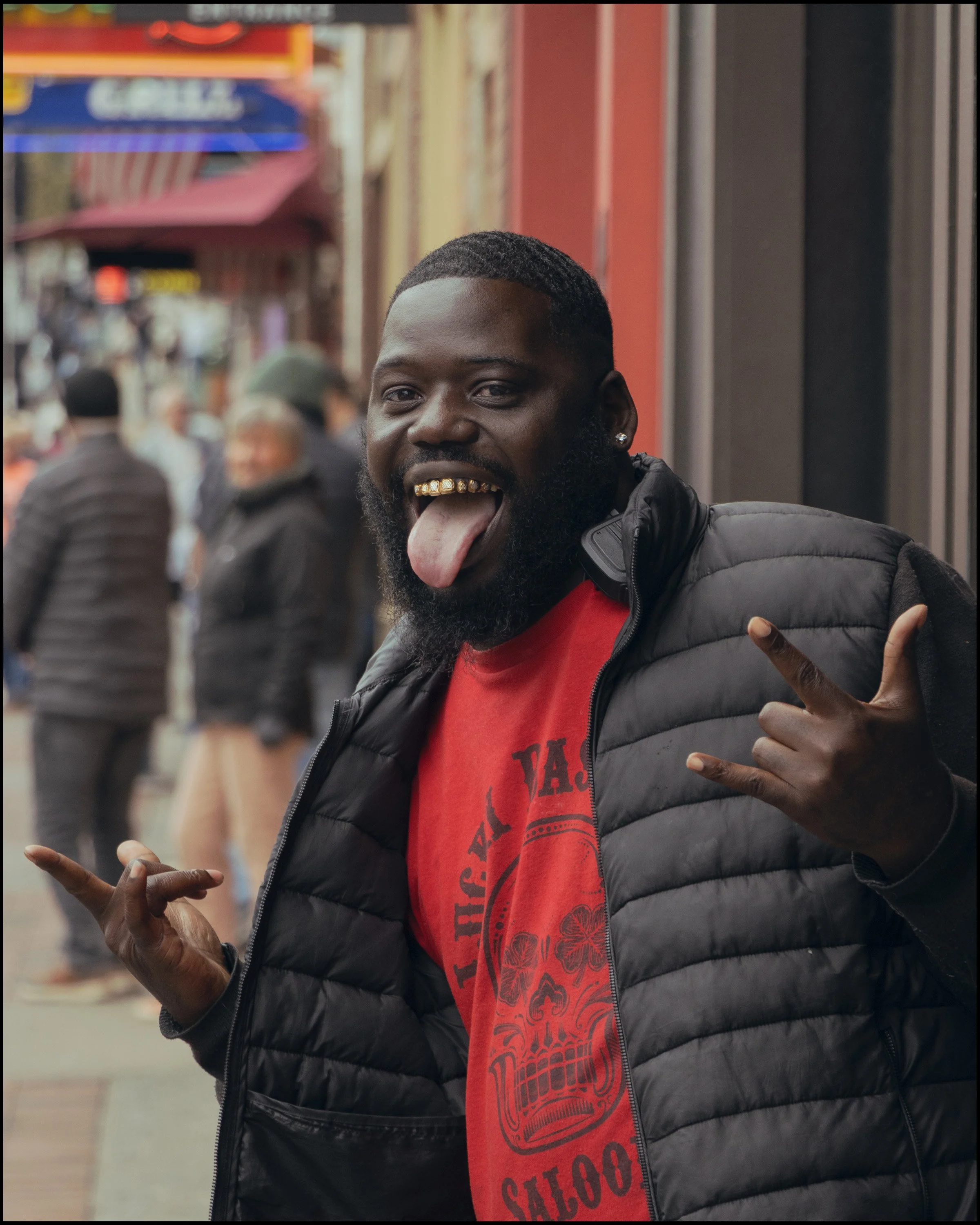 A young man with a beard and diamond earrings sticking his tongue out, wearing a red T-shirt and a black puffer jacket, posing with a peace sign on a city street with blurred pedestrians and storefronts in the background.