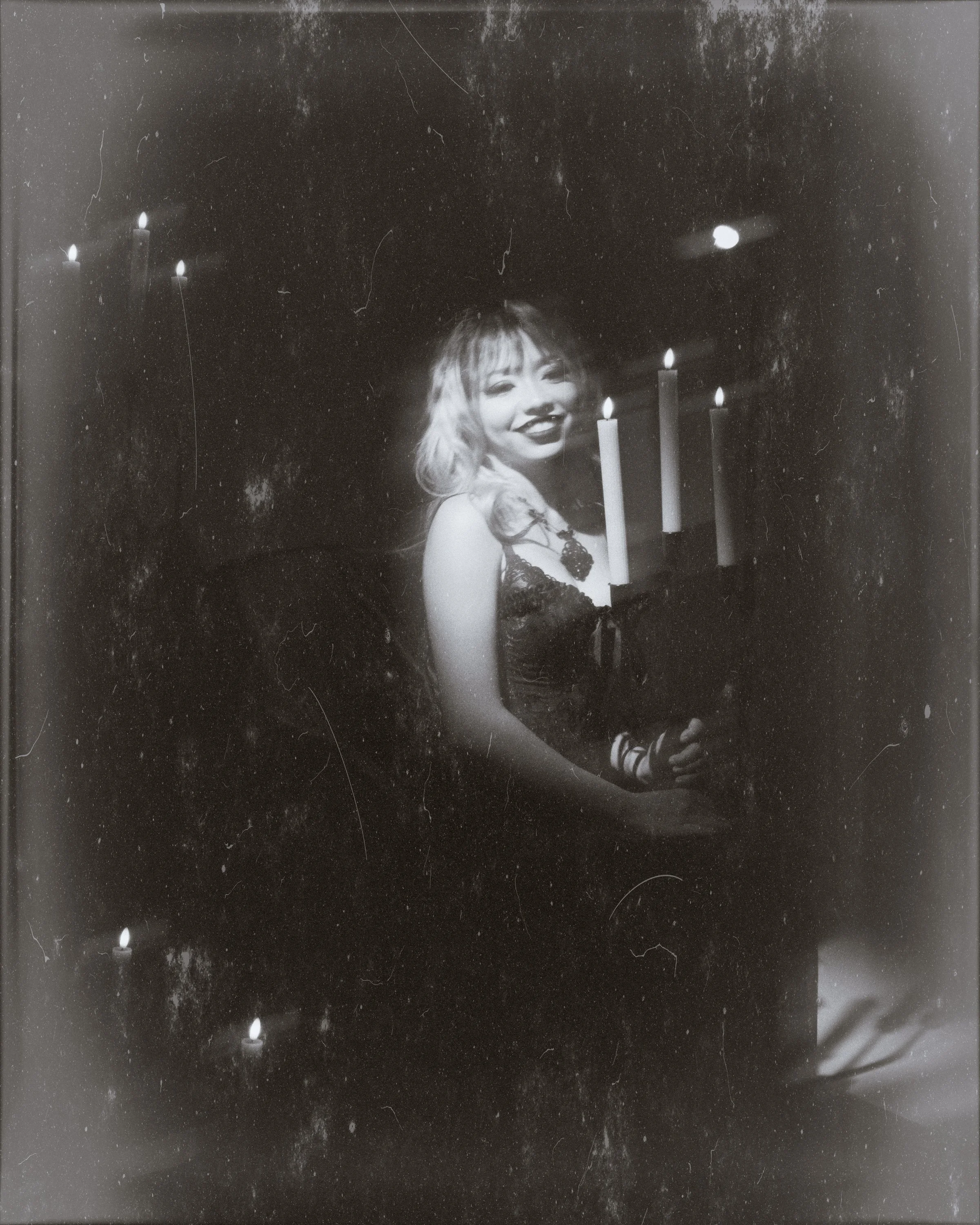 A black and white vintage photograph of a smiling woman with wavy hair, holding a candle, with a dark background and additional candles in the vicinity.