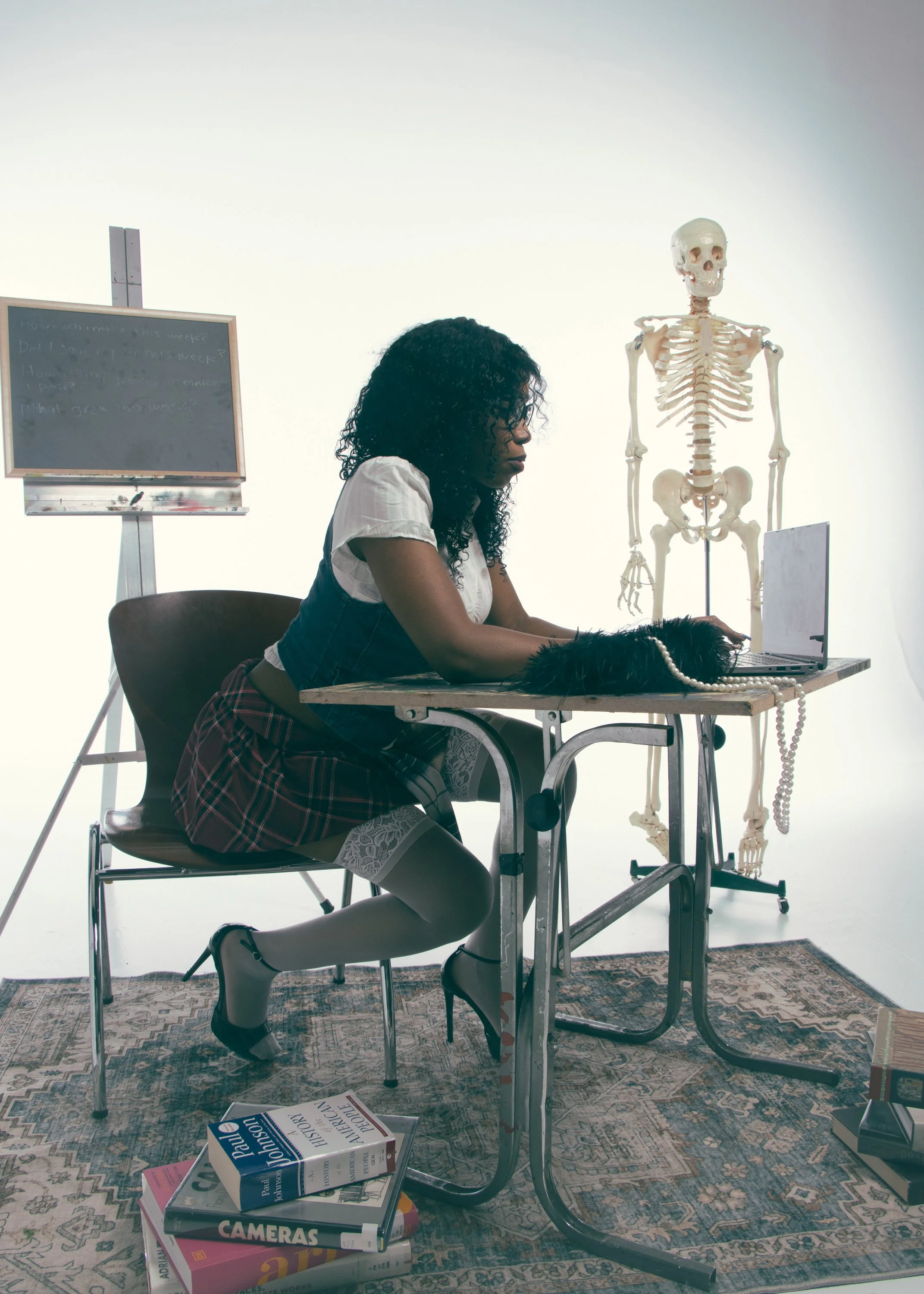 A woman with curly black hair, dressed in a white shirt, blue vest, and plaid skirt, sits at a wooden desk with her head bowed. On the desk are a skeleton model, a laptop, and a string of pearls. There are multiple books on history and cameras on the