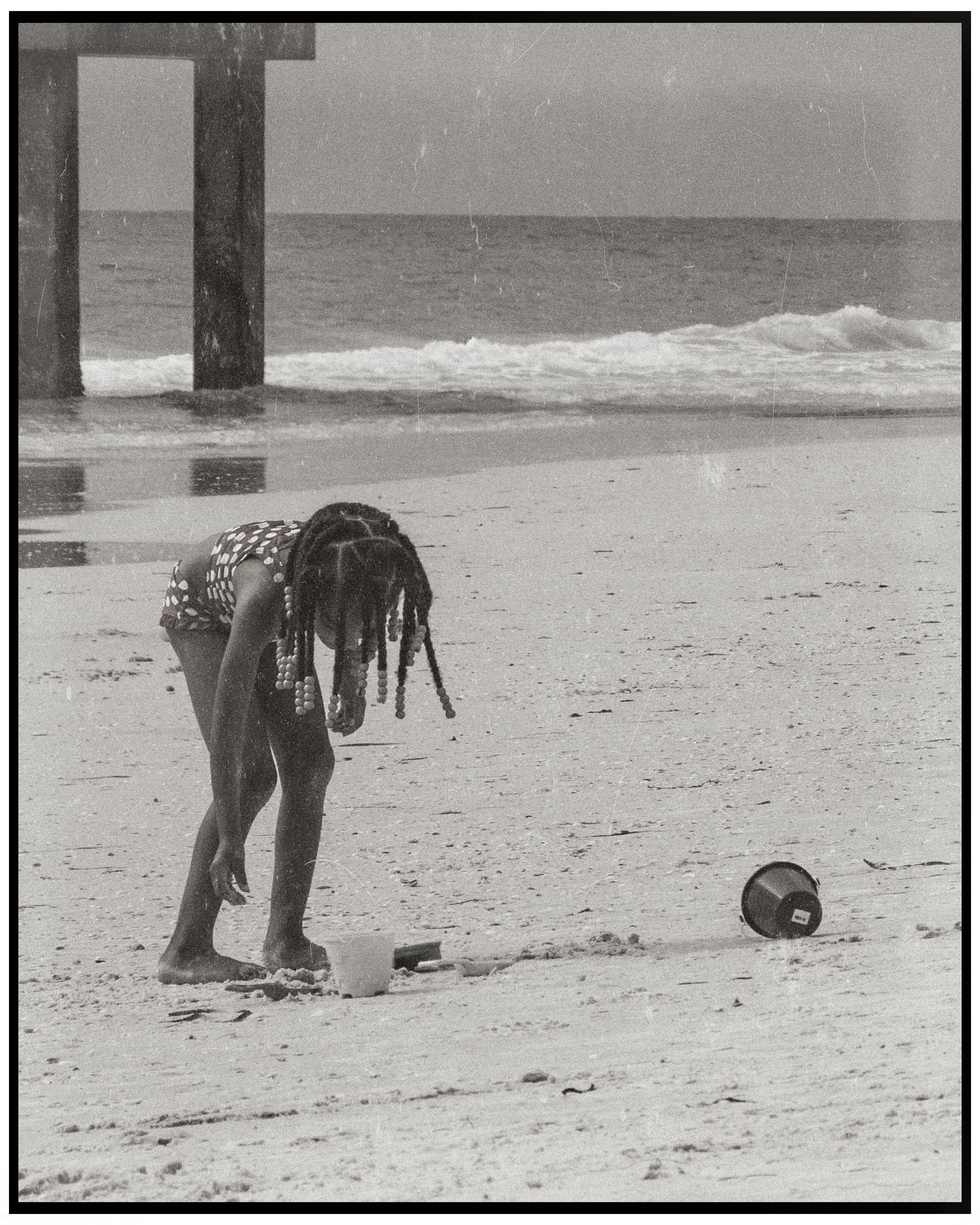 A black and white photo of a young girl with dreadlocks bending down on the beach, with a large wooden pier in the background and a beach ball on the sand nearby.