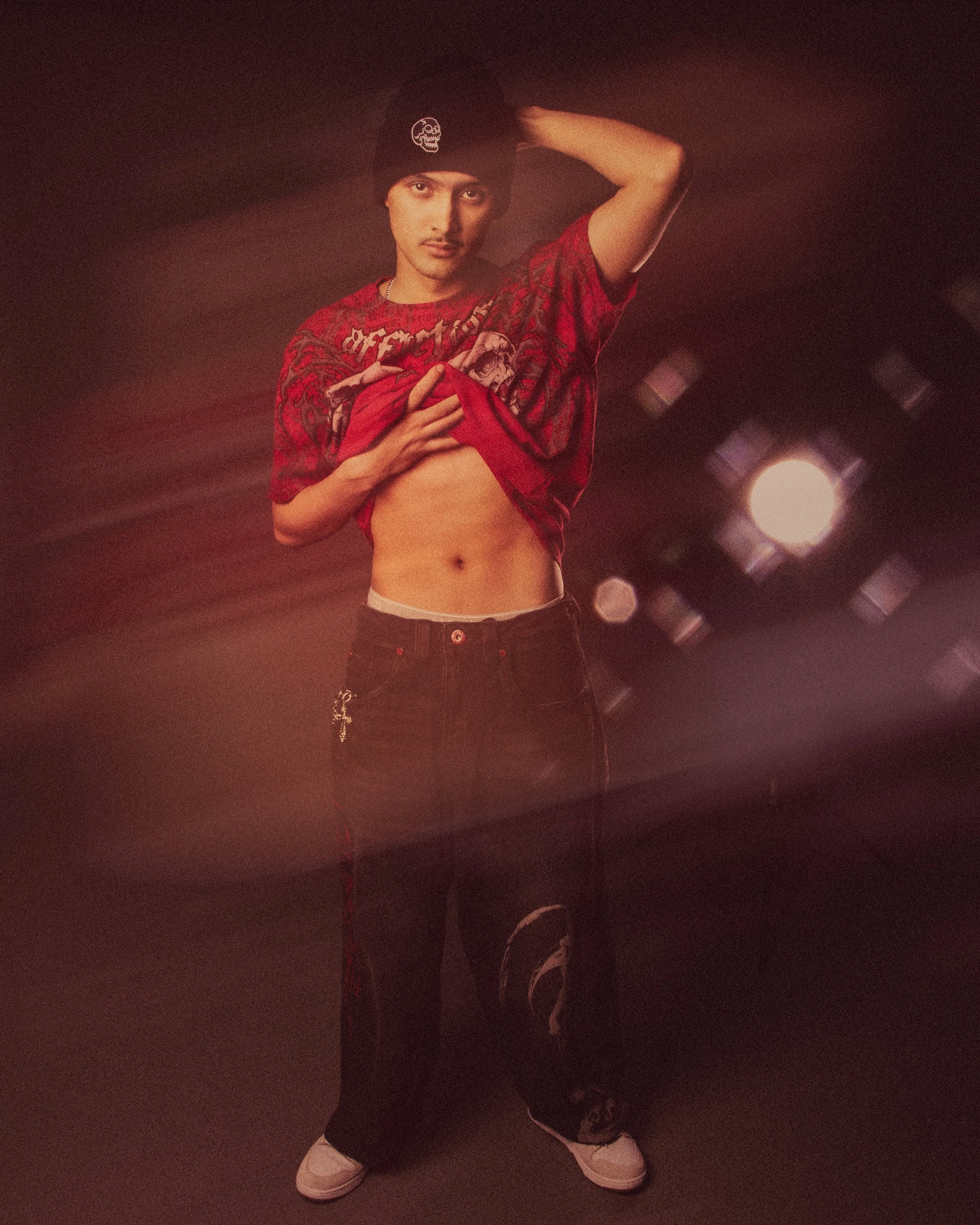 A young man wearing a black beanie with a skull emblem, red graphic T-shirt, black pants, and white sneakers, poses with one arm behind his head and the other hand lifting his shirt to reveal his abdomen, standing in a dimly lit room with overhead li