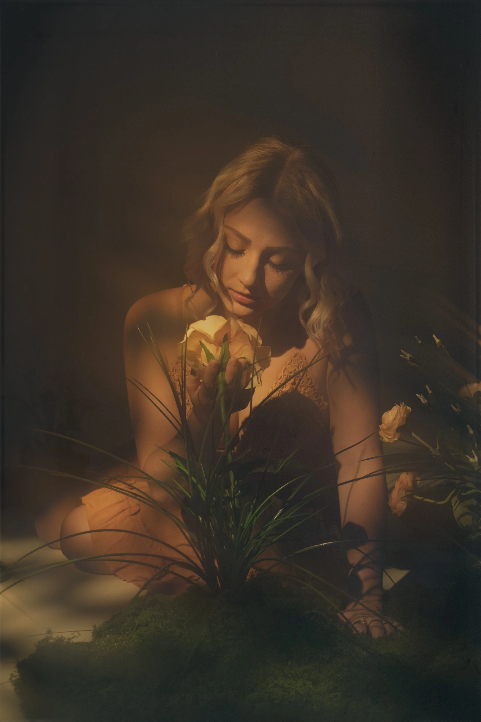 A woman with wavy blonde hair and closed eyes holding a bouquet of white flowers, surrounded by more flowers and greenery with a dark background.