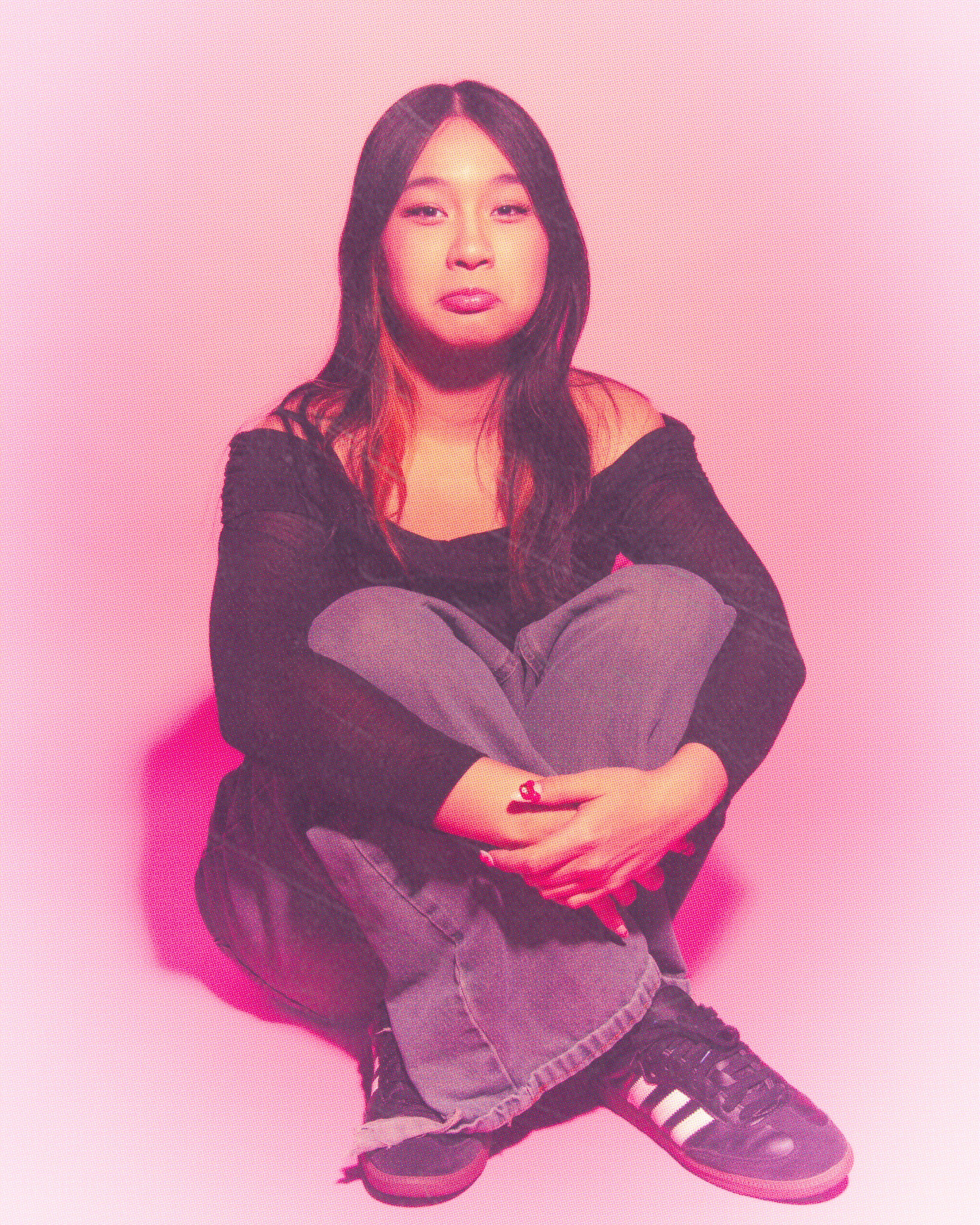 A woman with long dark hair sitting against a pink background, wearing a black off-the-shoulder top, ripped jeans, and black sneakers with white stripes.