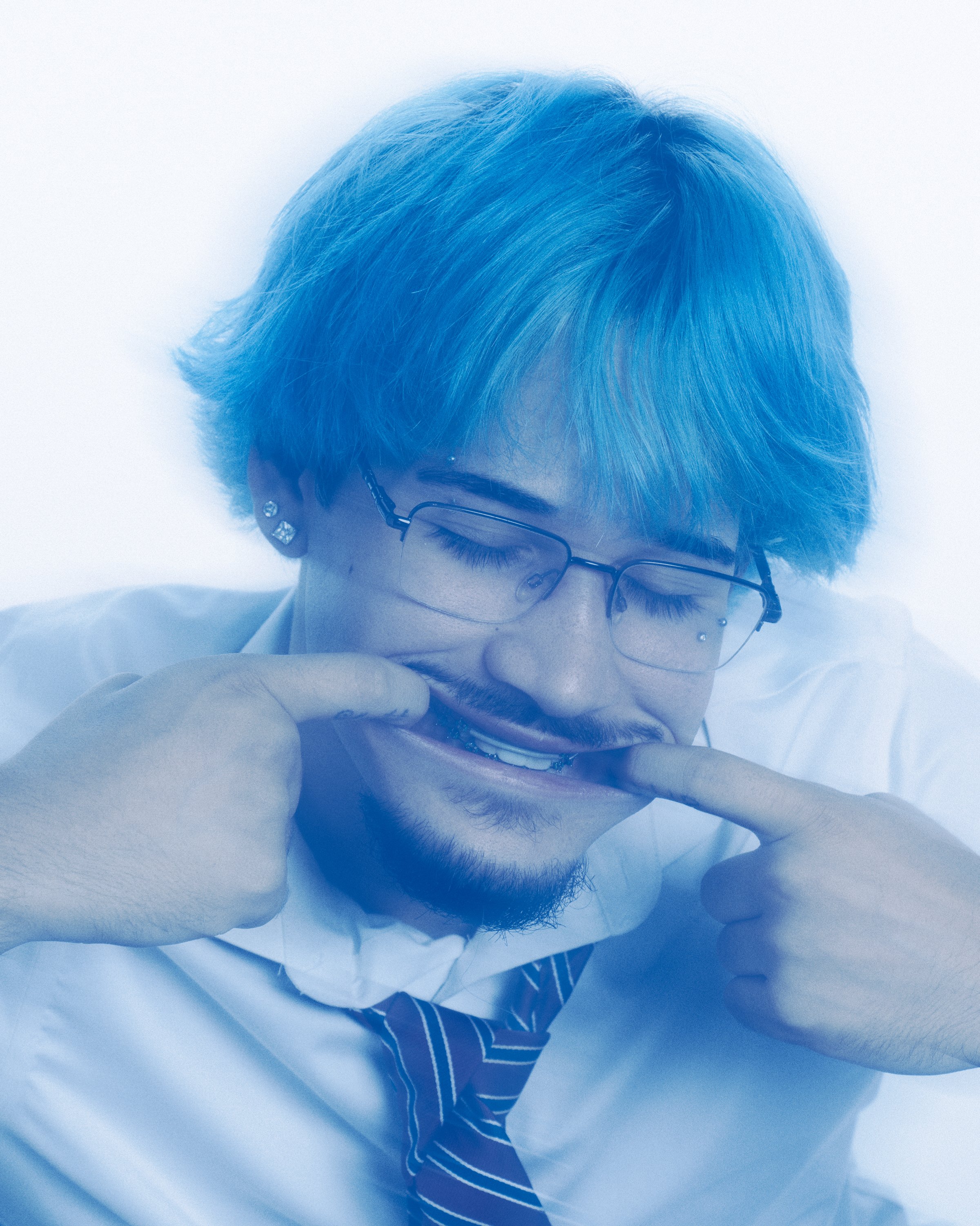 Person with short, blue hair, of Asian descent, wearing glasses, earrings, and a white shirt, resting their head on folded arms.