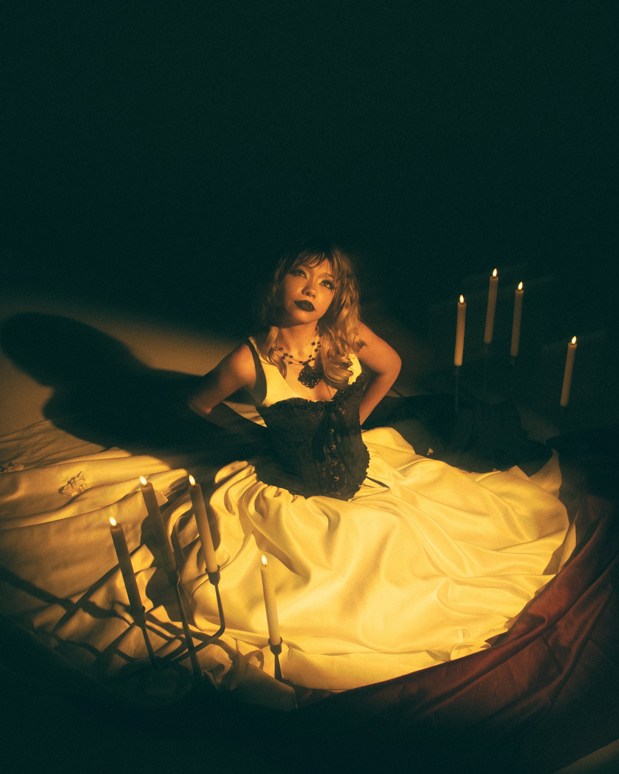 A woman with dark lipstick and curly hair sitting on a bed, surrounded by lit candles, in a dimly lit room.