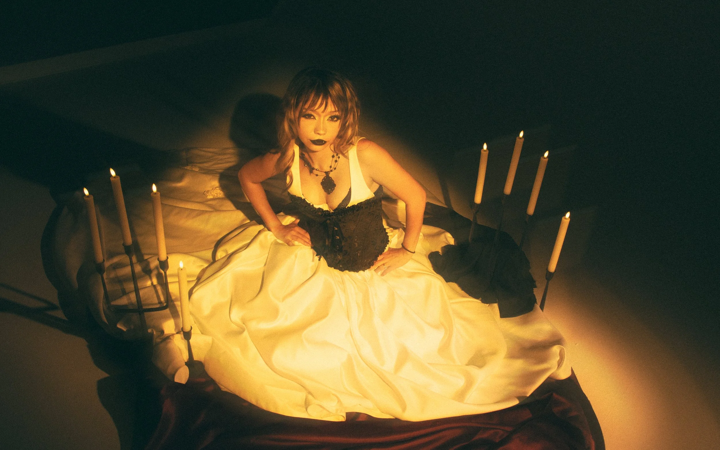 A woman sitting on a bed covered with a yellow satin sheet, surrounded by lit candles, with dark lipstick and layered jewelry.