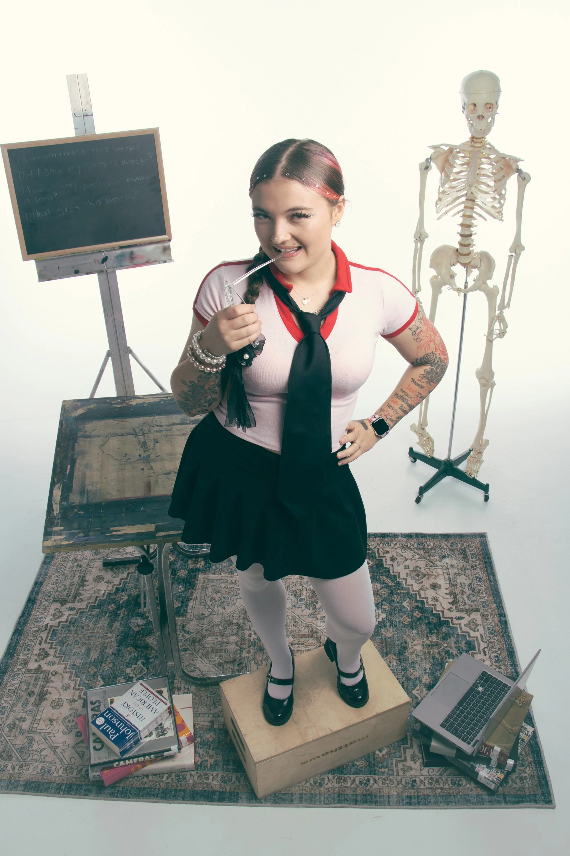 A young woman with tattoos, wearing a schoolgirl uniform with a white top, black tie, and black skirt, is standing on a wooden box surrounded by books, a laptop, and a blackboard in a studio setting. She is holding a glass straw near her mouth with o