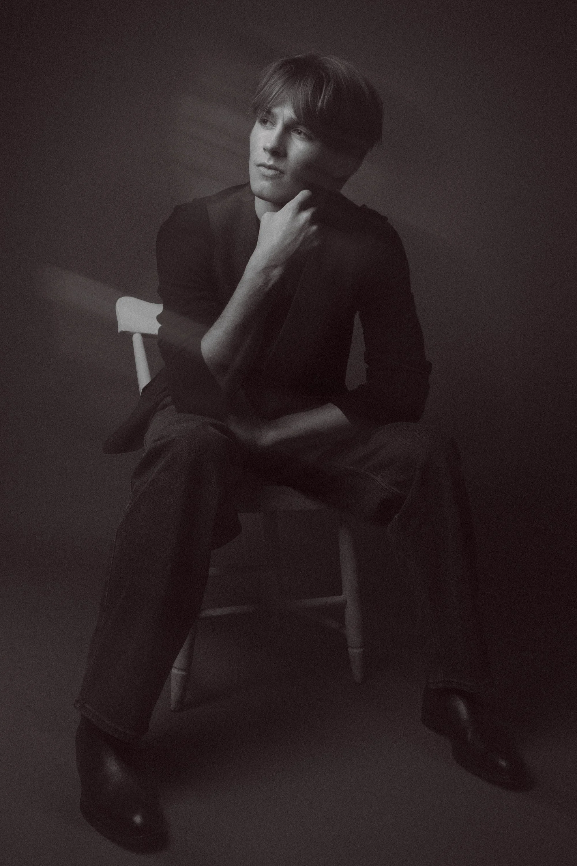 Black and white photo of a young man sitting on a chair, resting his chin on his hand, with a thoughtful expression. He has medium-length hair and is dressed in dark clothing. The background is plain and dark.
