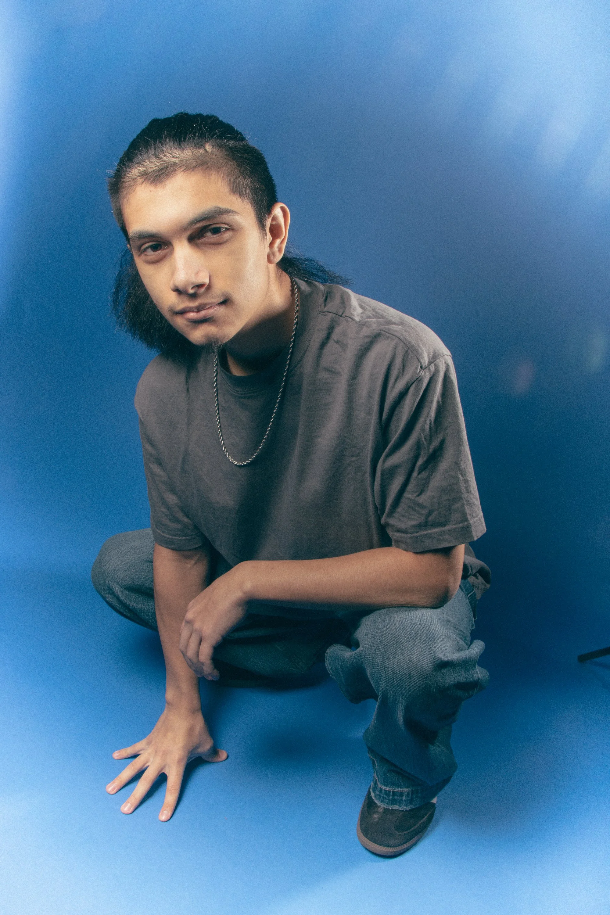 A young man with long dark hair partially shaved on the sides, wearing a gray T-shirt, jeans, and sneakers, crouching on a blue background, looking at the camera with a slight smile, with one hand on the ground and the other on his knee.