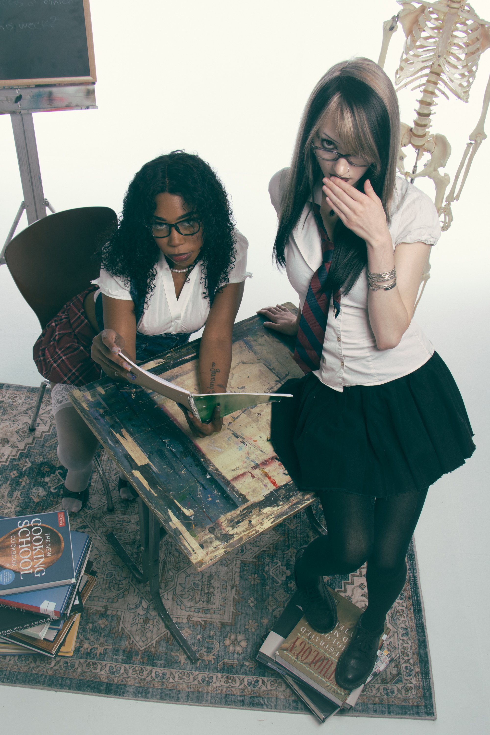 Two women in school uniforms with glasses, one with curly black hair and the other with straight brown hair, looking at a book on a cluttered table in an art studio, with a human skeleton model in the background.