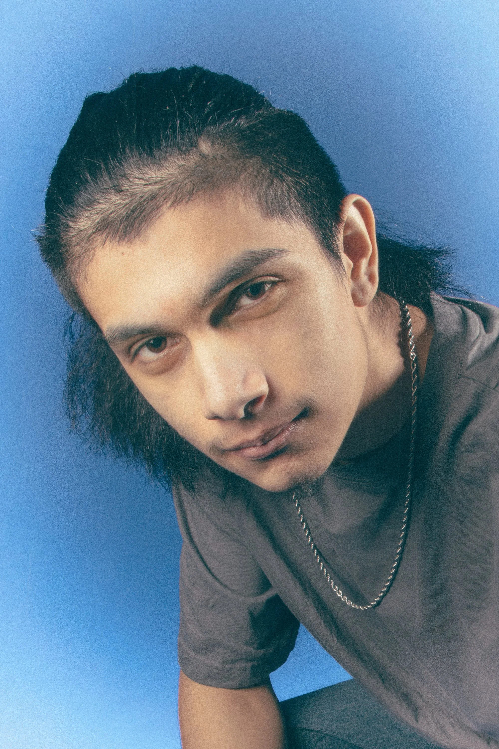 Close-up of a young man with dark hair and a mustache, wearing a gray T-shirt and a chain necklace, leaning forward against a blue background.