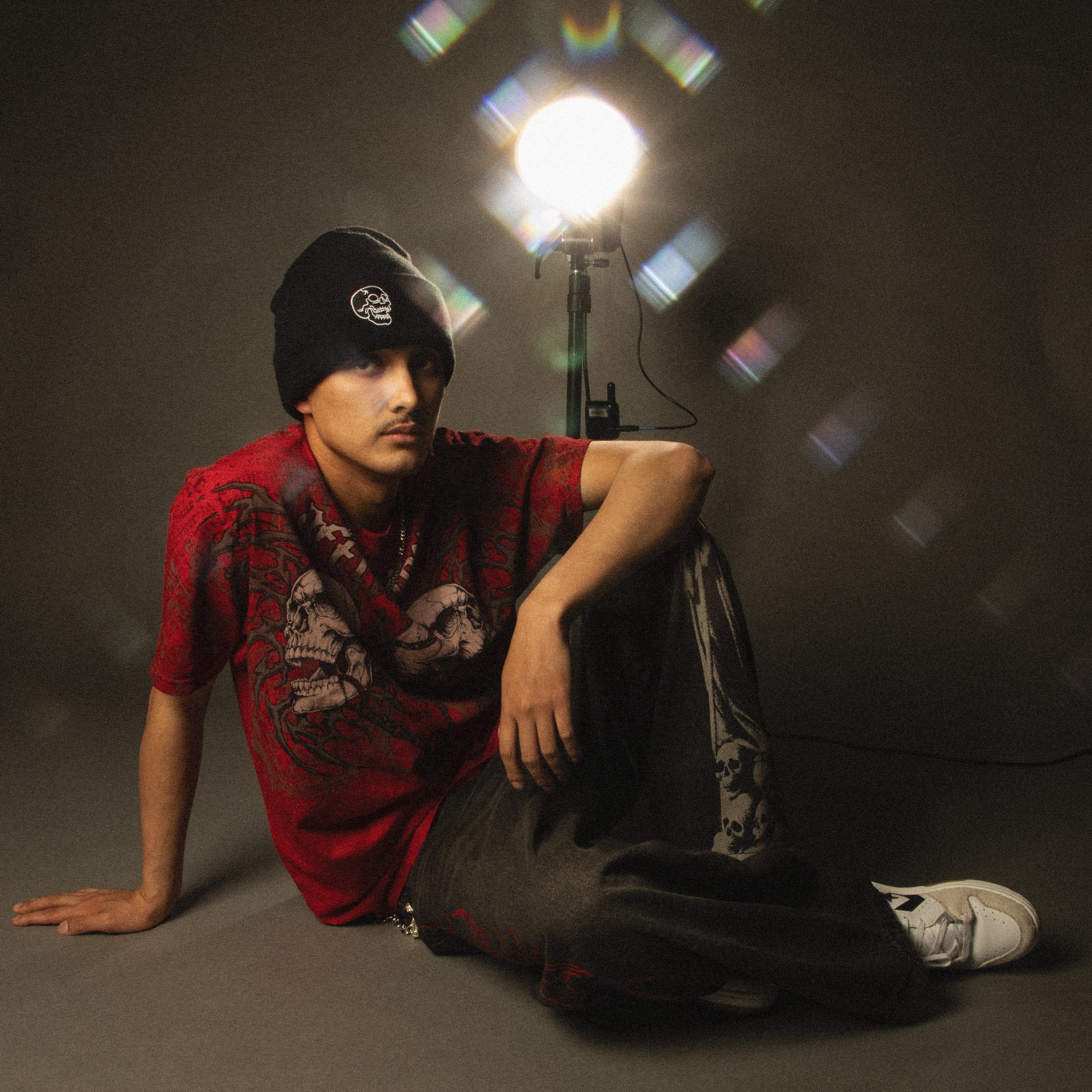 A person wearing a black beanie with a skull logo, red shirt with skull design, and sneakers, is lying on their side on a dark surface with a bright studio light behind them, creating refractions in the lens.