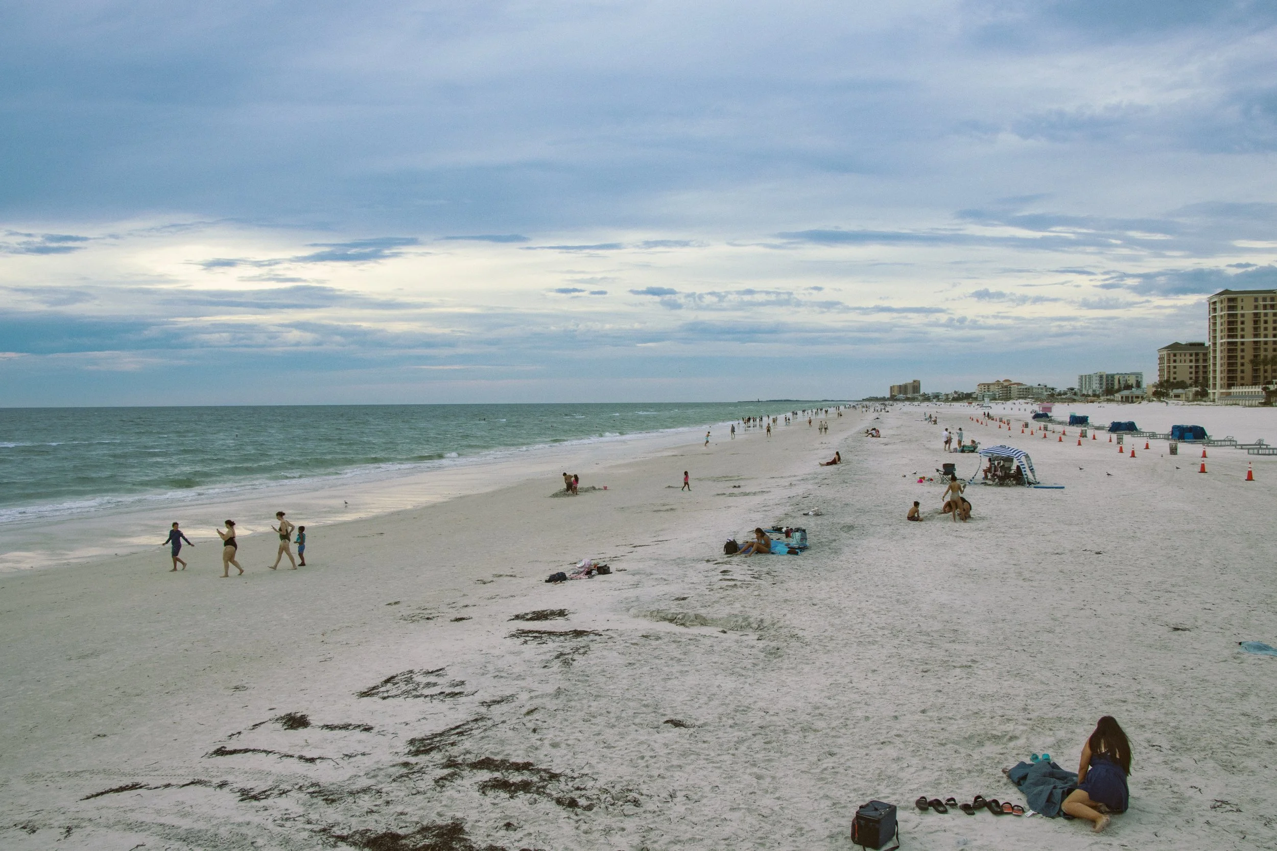 A beach scene with people walking, sitting, and relaxing on white sand under a partly cloudy sky. High-rise buildings are visible along the shoreline in the background.