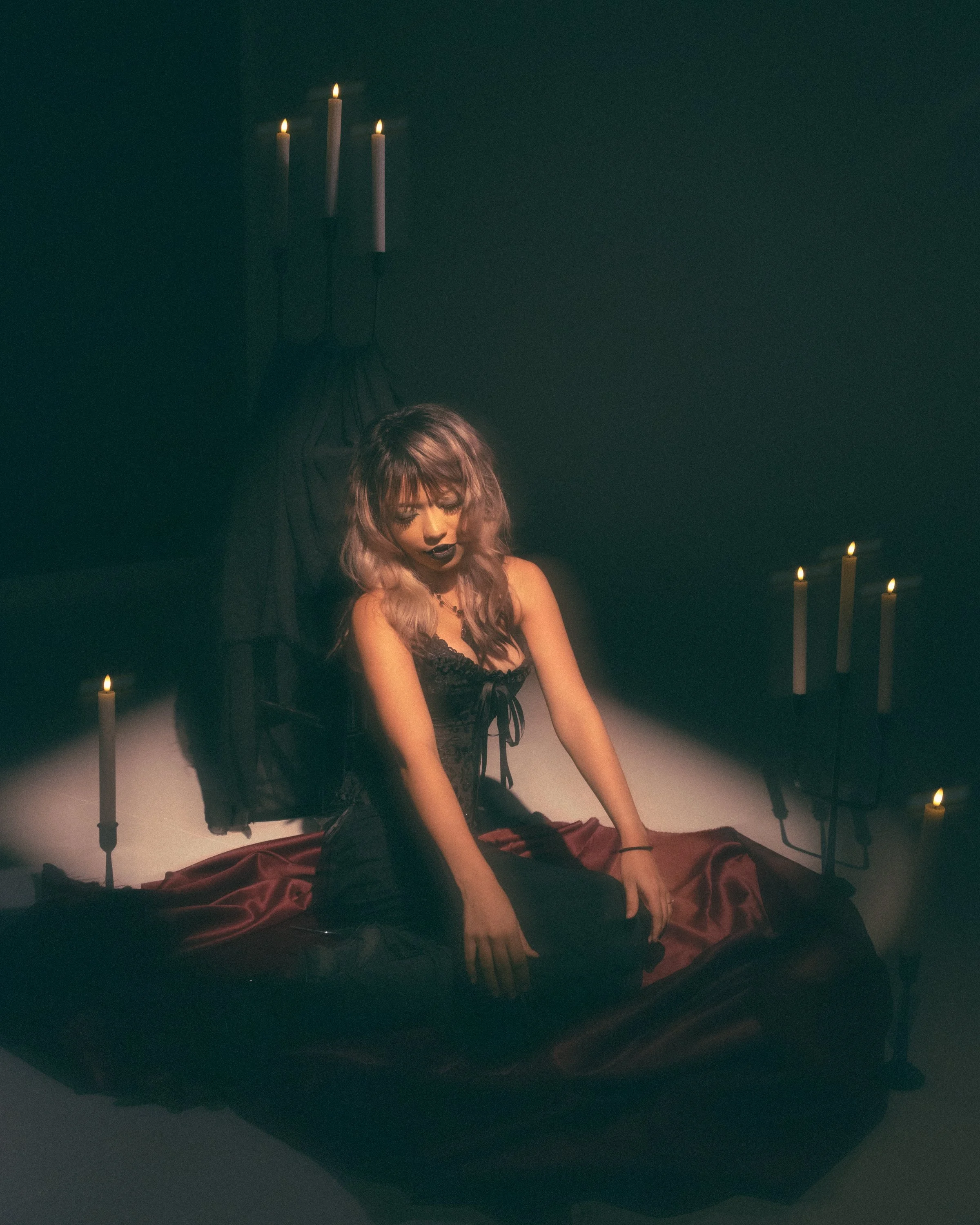 A woman with long, wavy, pastel-colored hair seated on the floor, surrounded by lit candles in a dark room with a black wall. She is wearing black lingerie and appears to be deep in thought or meditation.