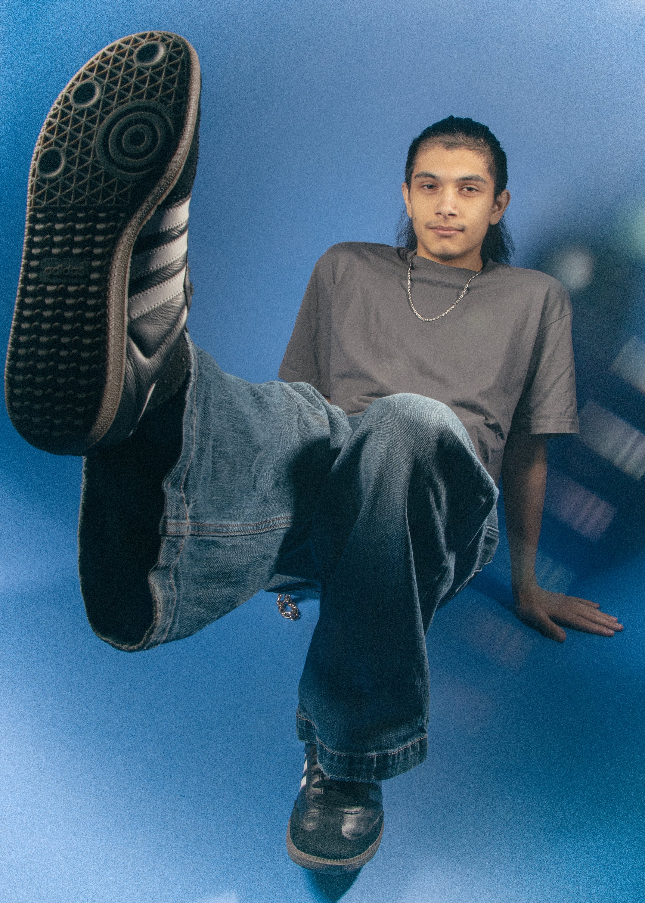 Young man with long hair and a chain necklace sitting with his leg extended toward the camera, wearing jeans, a brown t-shirt, and black shoes, against a blue background.
