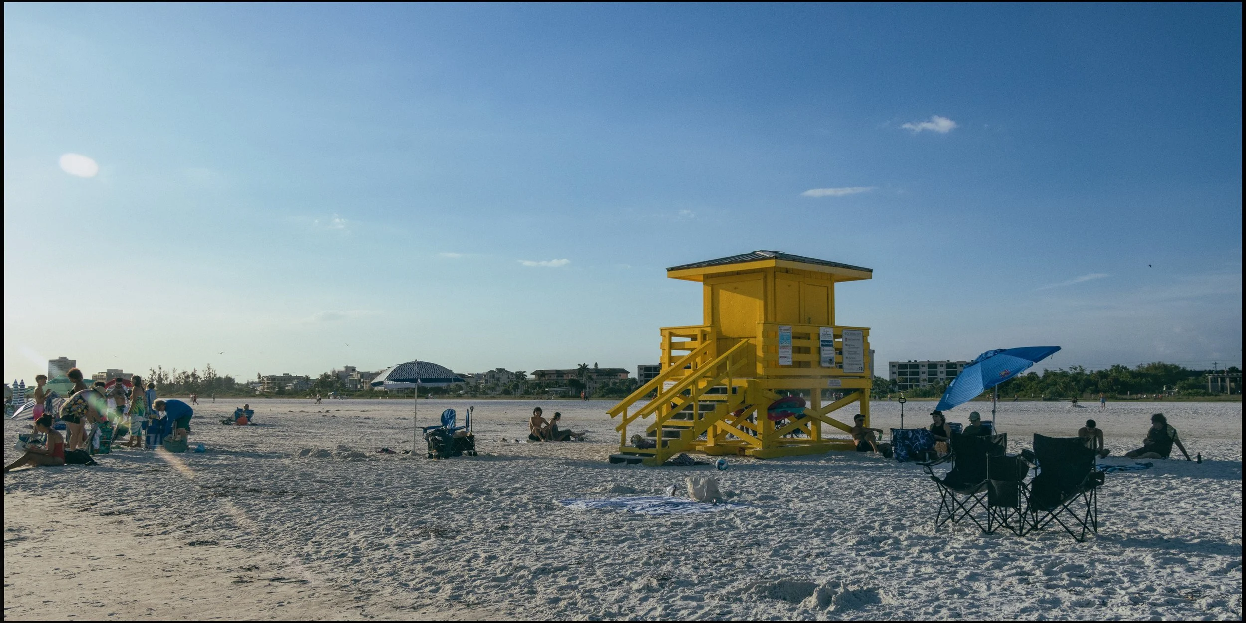 People relaxing on a beach with blue umbrellas, a yellow lifeguard tower, and buildings in the background on a sunny day.