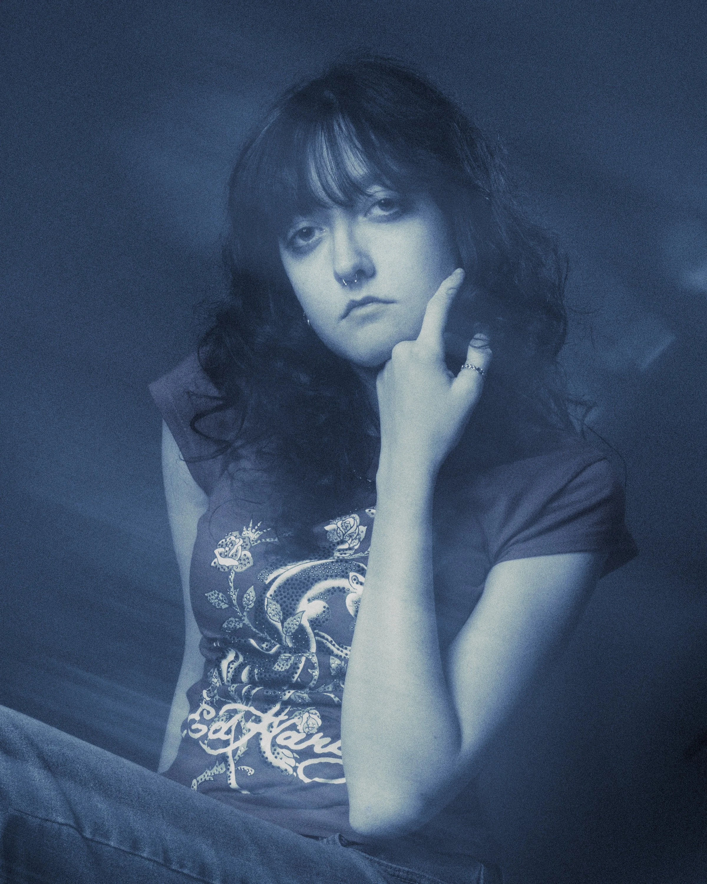 A young woman with dark, curly hair, wearing a nose ring and rings on her fingers, resting her face on her hand, dressed in a dark T-shirt with a graphic design, against a dark background.