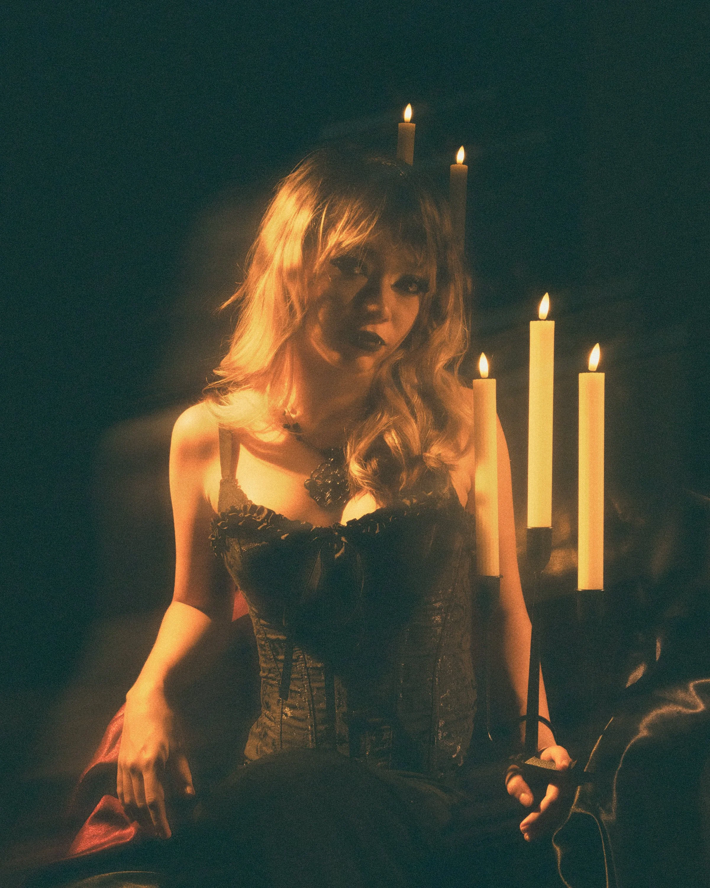A woman with wavy blonde hair and dark lipstick wearing black lingerie, surrounded by lit candles in a dark setting.