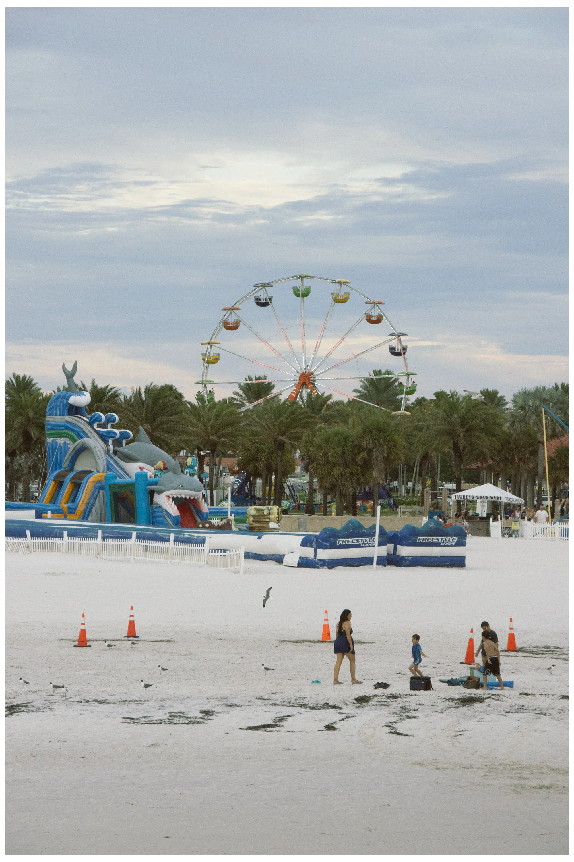 A beach with a water park featuring an inflatable shark slide and various water rides, surrounded by palm trees. In the background, there is a large Ferris wheel and amusement park rides, with a cloudy sky overhead. Several people are walking and chi