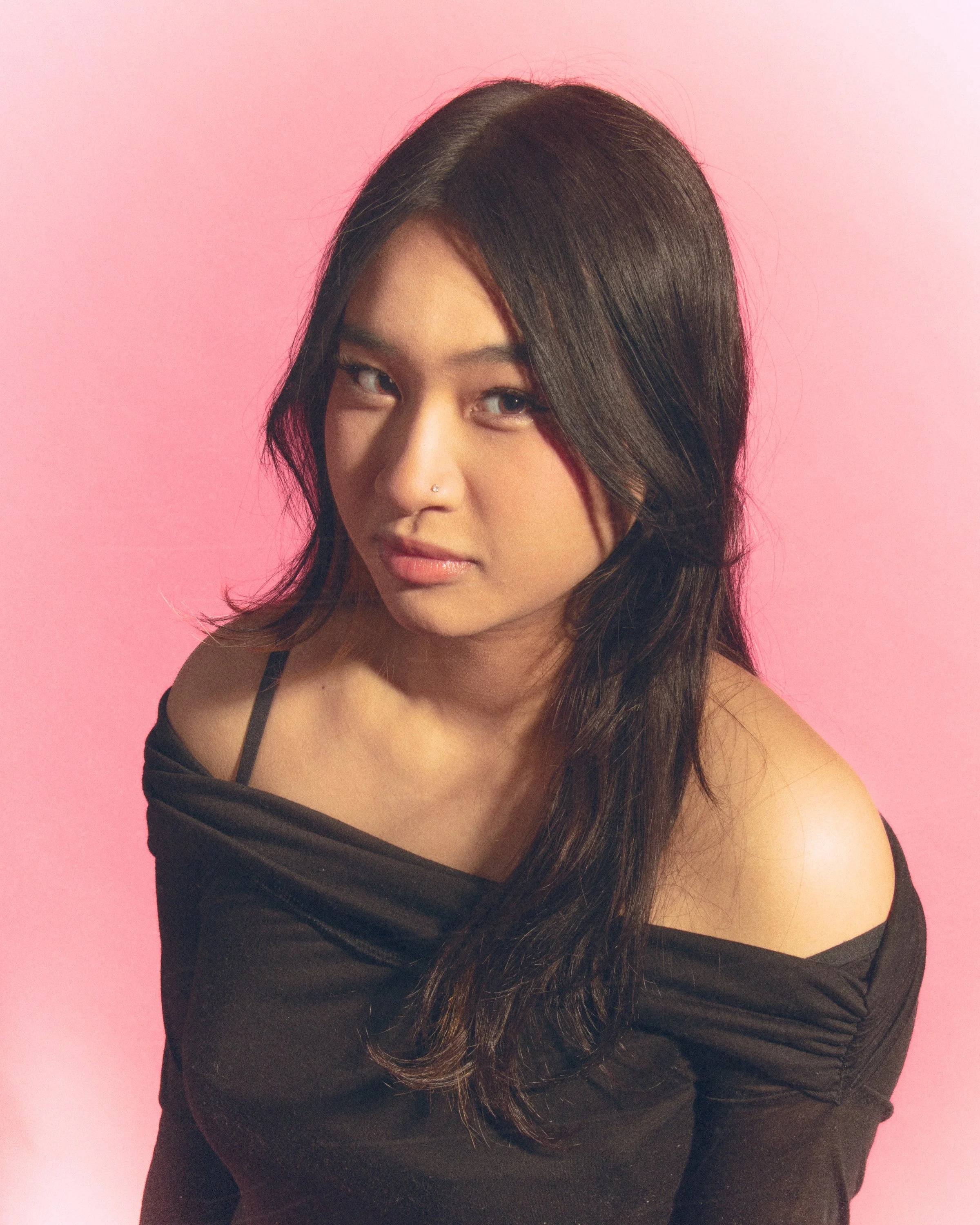 Close-up of a young woman with dark brown hair, wearing a black off-shoulder top, against a pink background.