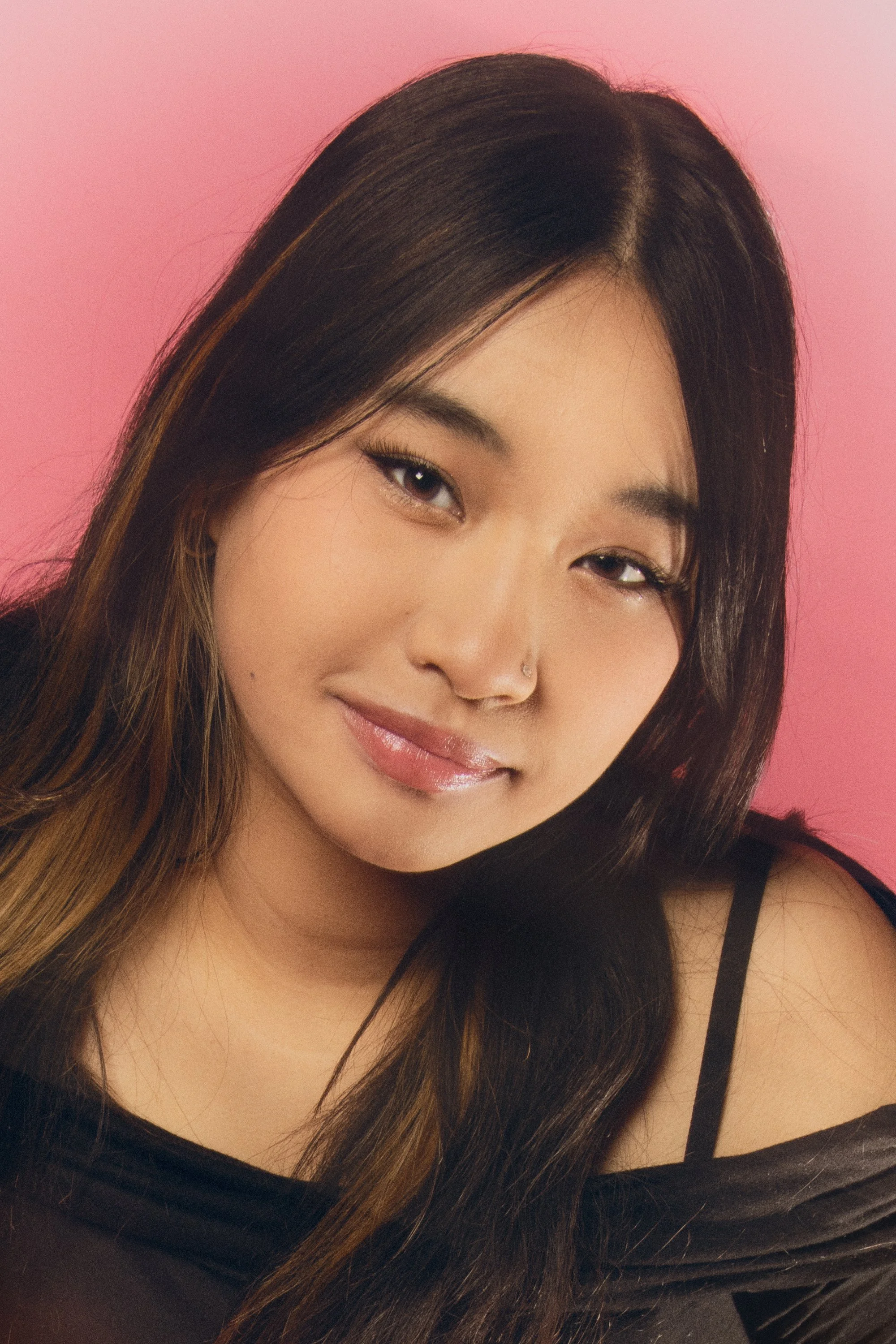 Close-up of a young Asian woman with long, dark hair against a pink background. She is smiling subtly and wearing a black top with thin straps.