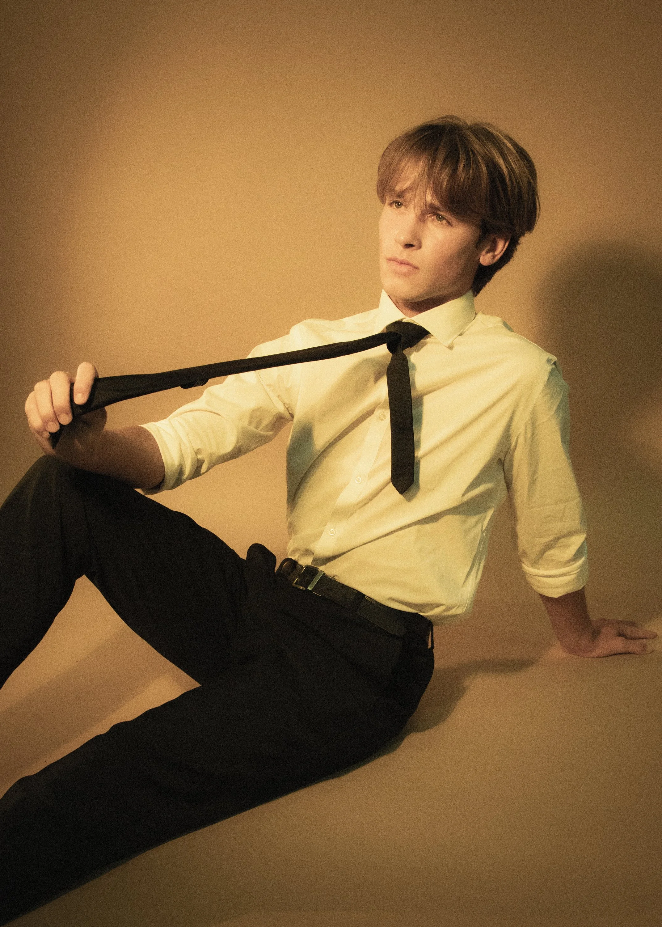 Young man with brown hair, wearing a yellow shirt and black tie, seated on the floor against a beige wall, holding a black belt.