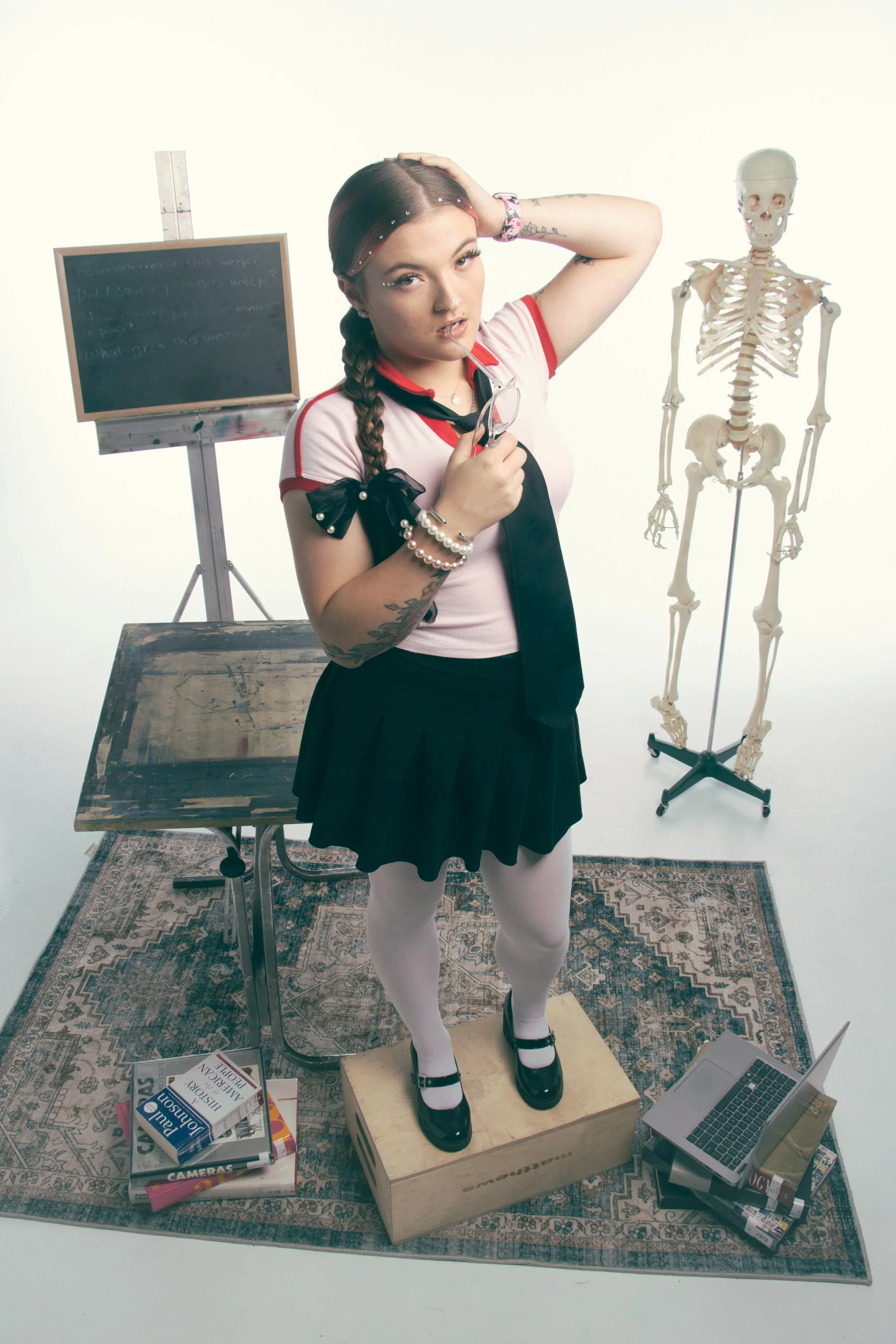 A young woman with braided hair and tattoos stands on a wooden box, holding glasses and a straw in her mouth, in a studio setting with skeleton model, chalkboard, magazines, laptop, and art supplies around her.