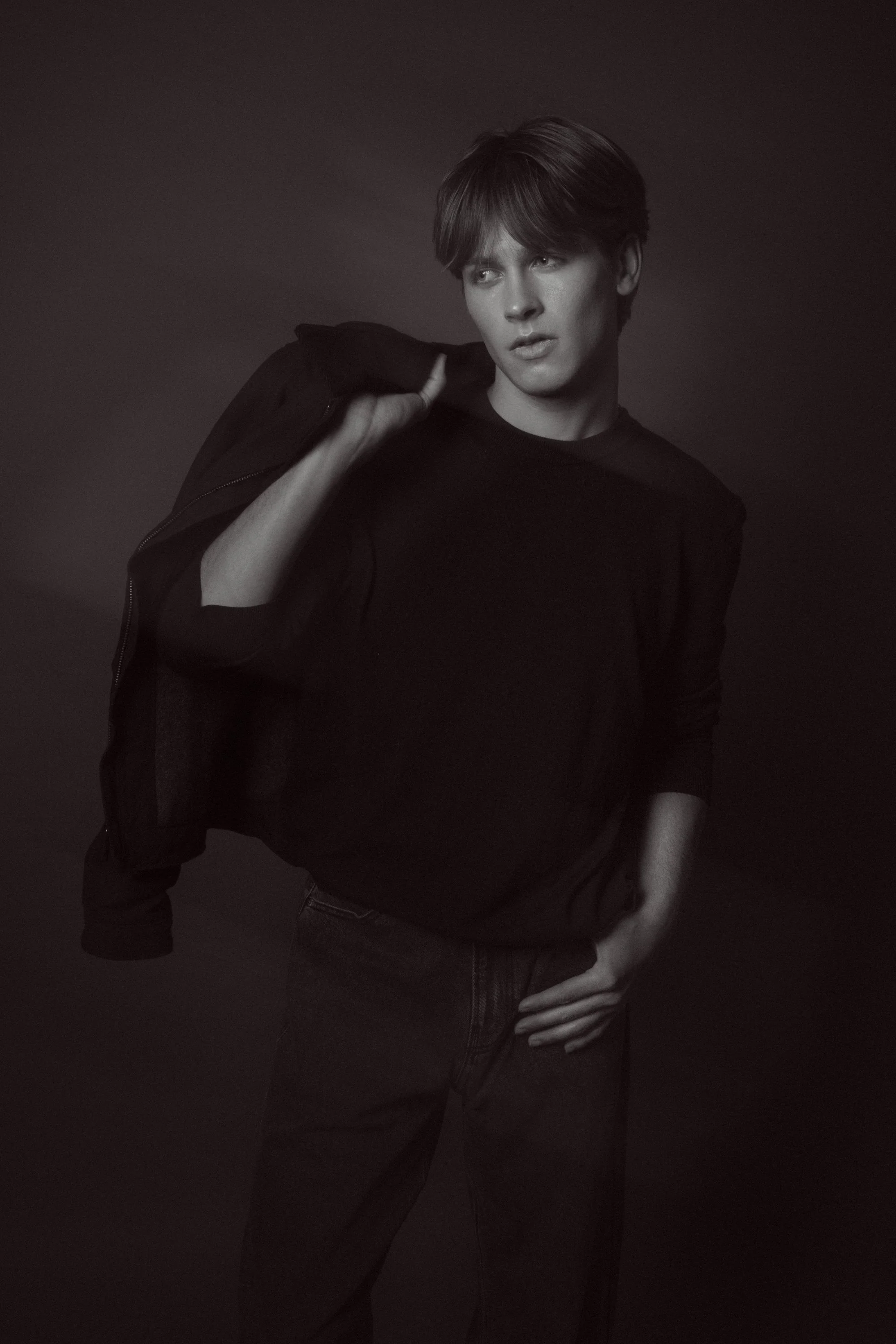 A black-and-white photo of a young man with short hair, wearing a dark sweater, holding a jacket over his shoulder, with one hand on his hip, standing against a dark background.