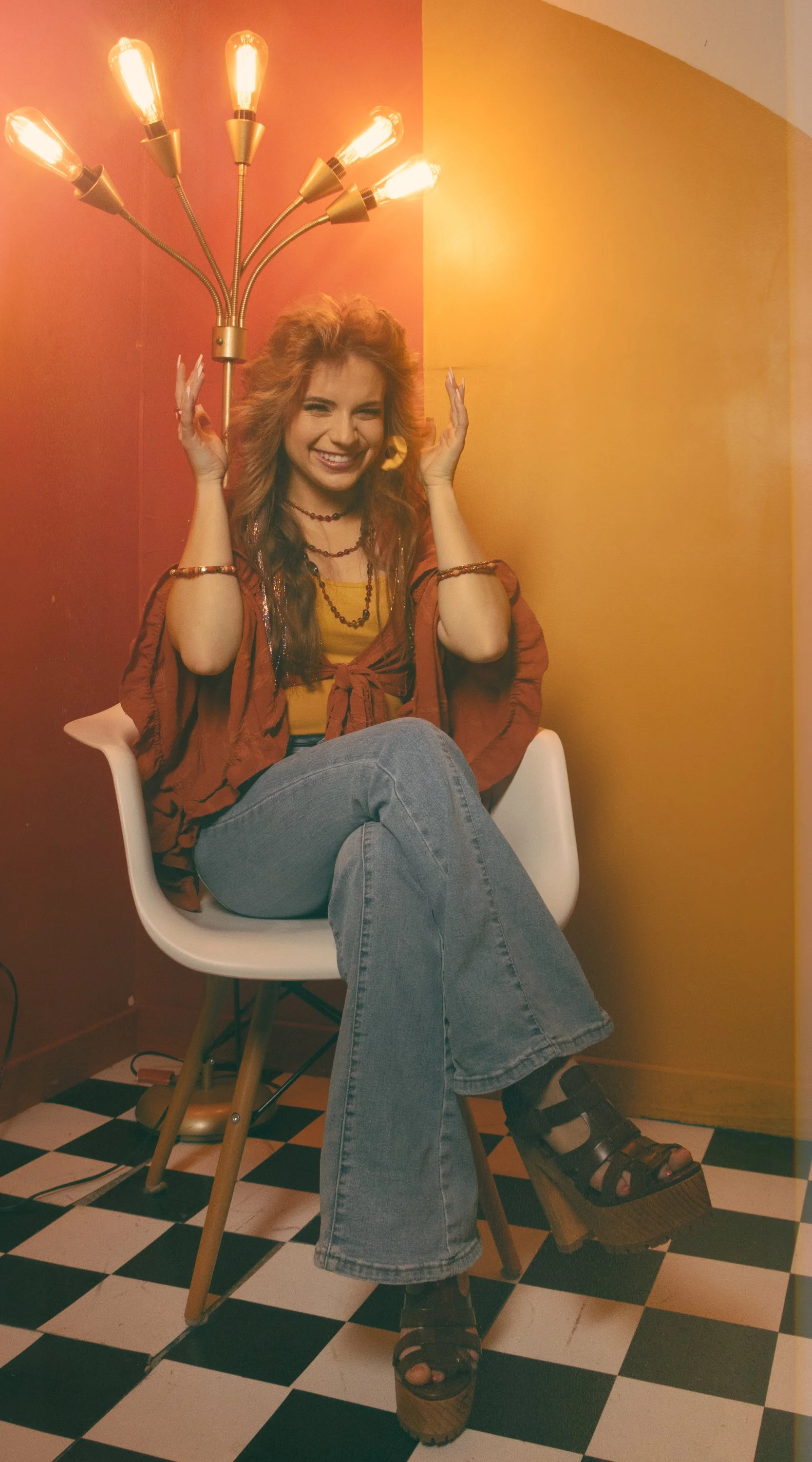 A young woman sitting in a white chair in a checkerboard black and white floor room, smiling and winking, holding a multi-armed brass and gold floor lamp above her head, with yellow and orange walls.