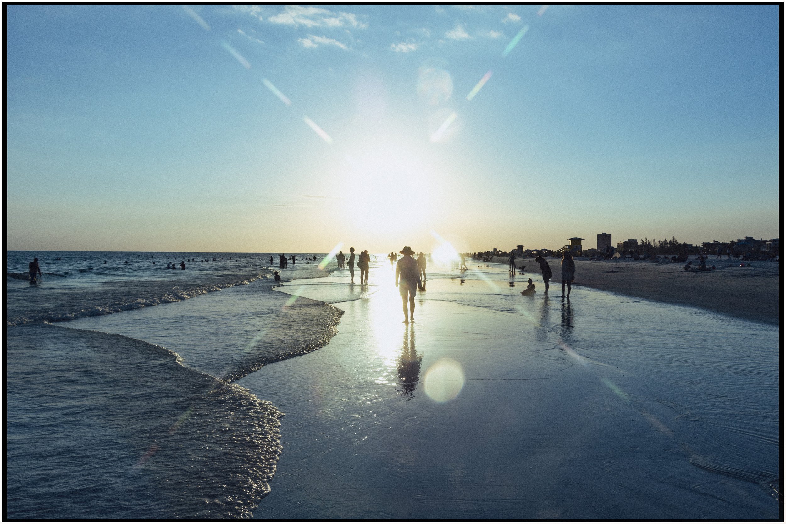 Sunset over a crowded beach with people walking, swimming, and relaxing along the shoreline and in the water.