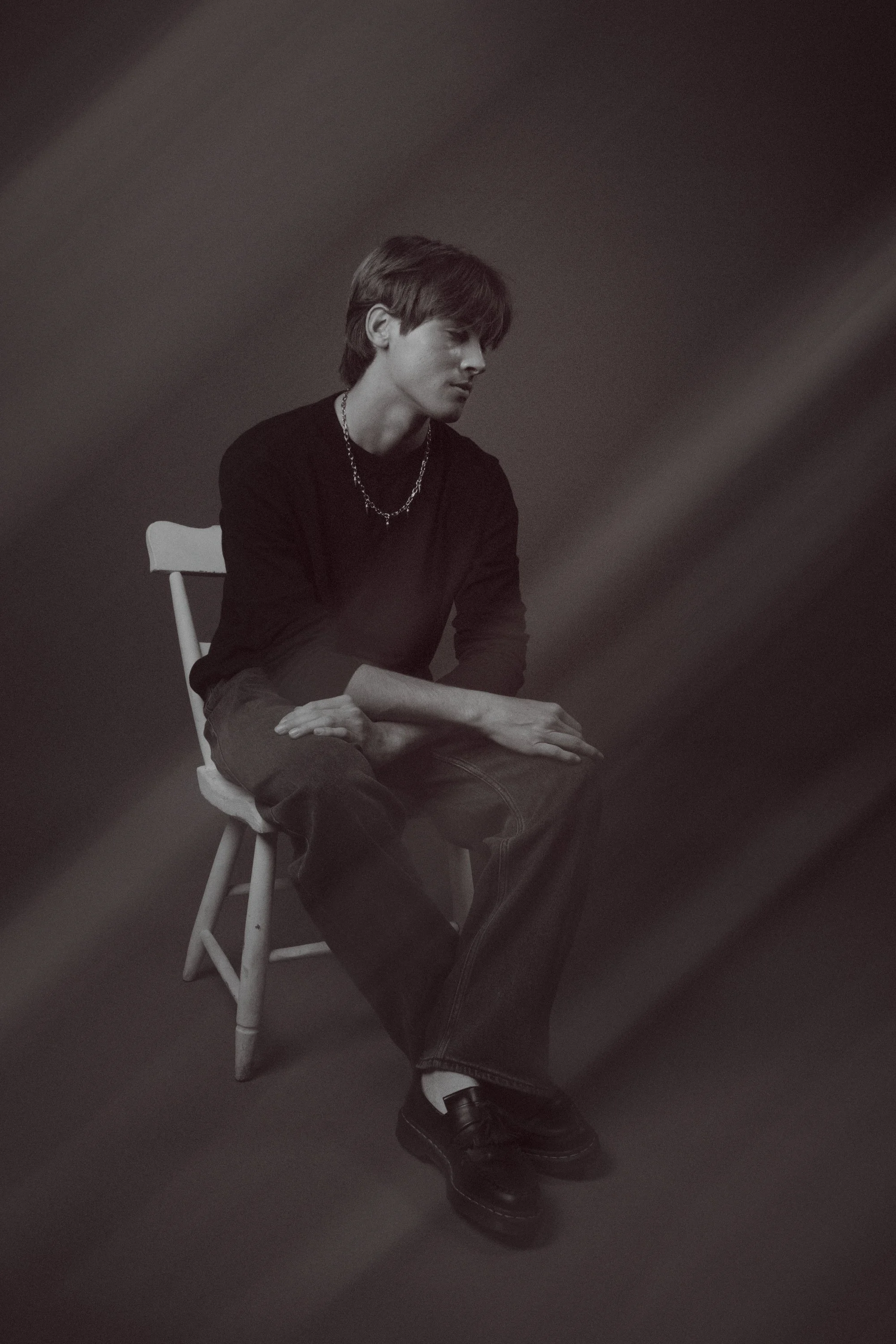 Black and white photo of a young man sitting on a white chair, looking down with a pensive expression, wearing a black shirt, chain necklace, jeans, and black boots, against a dark background.