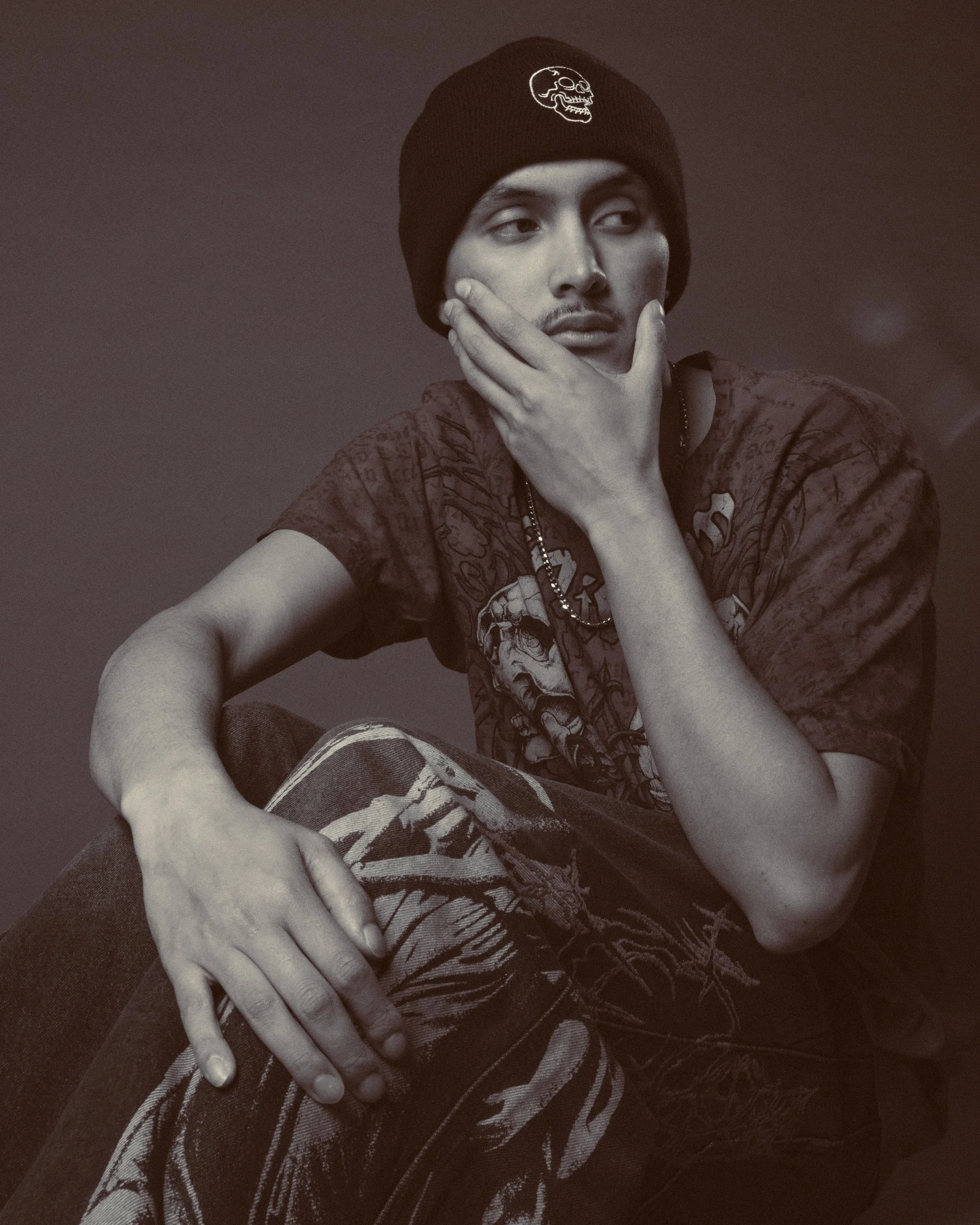 Black and white photo of a person wearing a beanie with a skull emblem, resting their chin on their hand, wearing a graphic T-shirt and sitting on a zebra-patterned blanket.